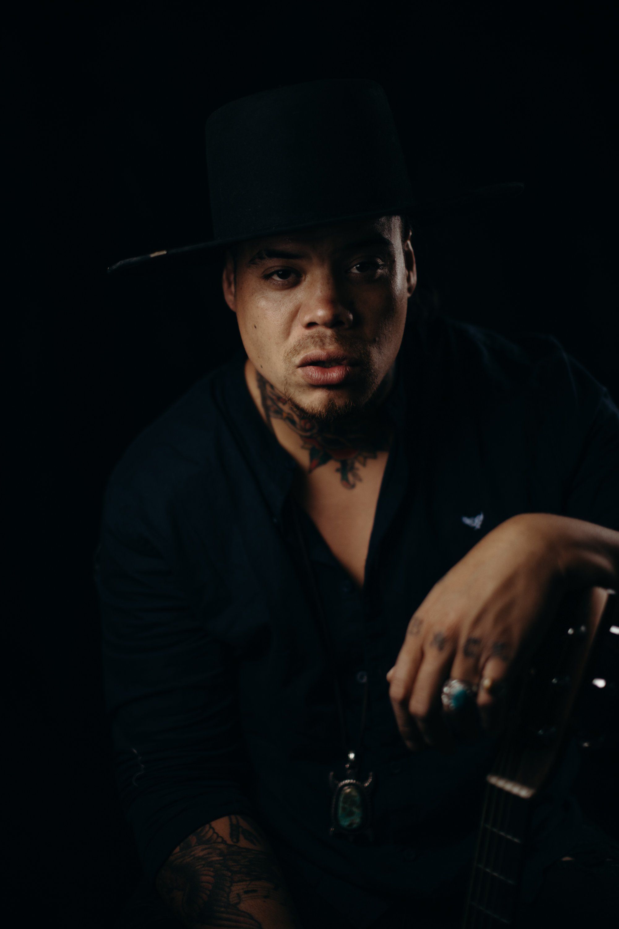 Man in black shirt and wide-brimmed hat gazes toward camera against dark background, dramatic side lighting emphasizing facial features and contemplative mood