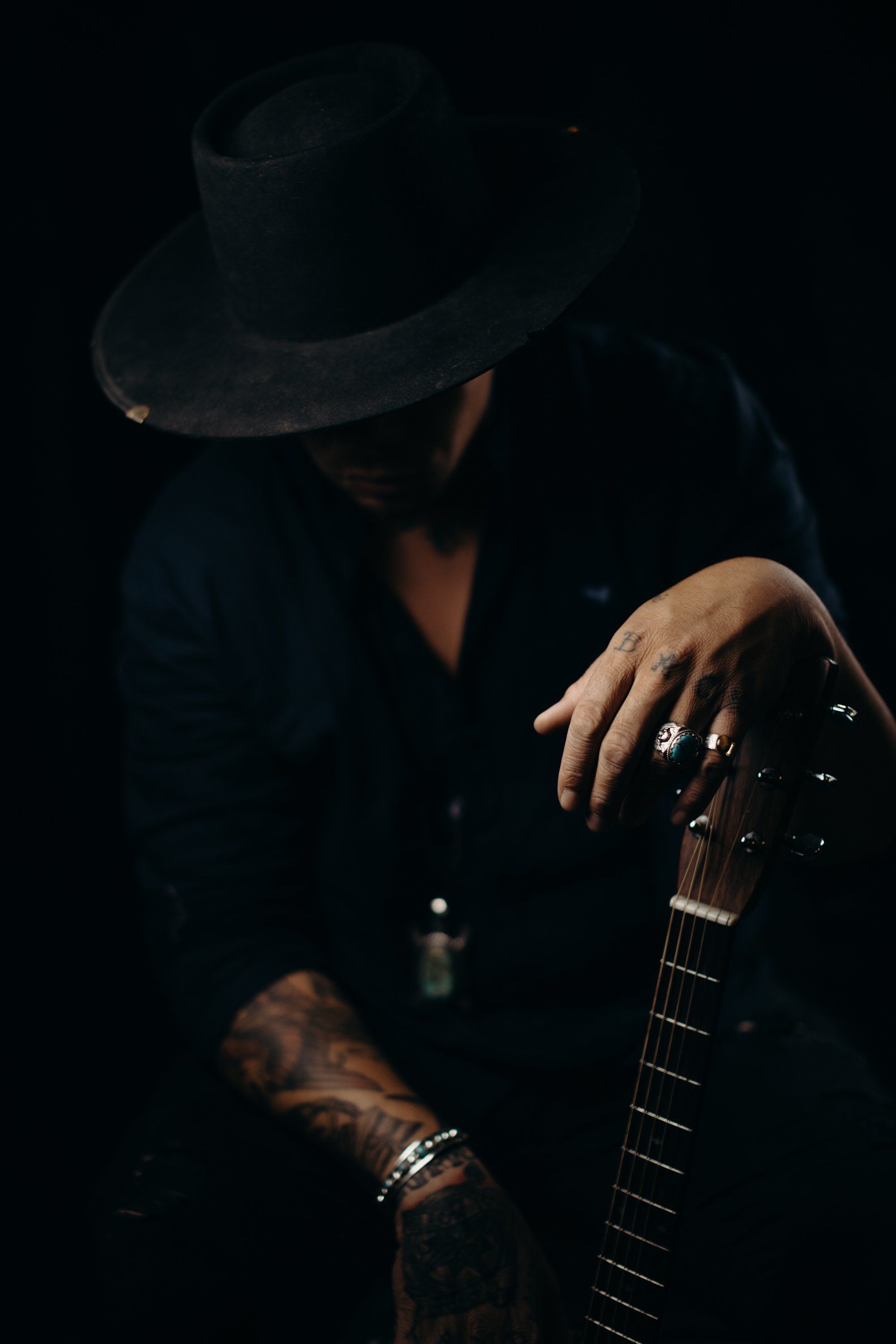 Musician in black hat and jacket holds electric guitar against dark background, rings glinting on fingers, moody studio portrait