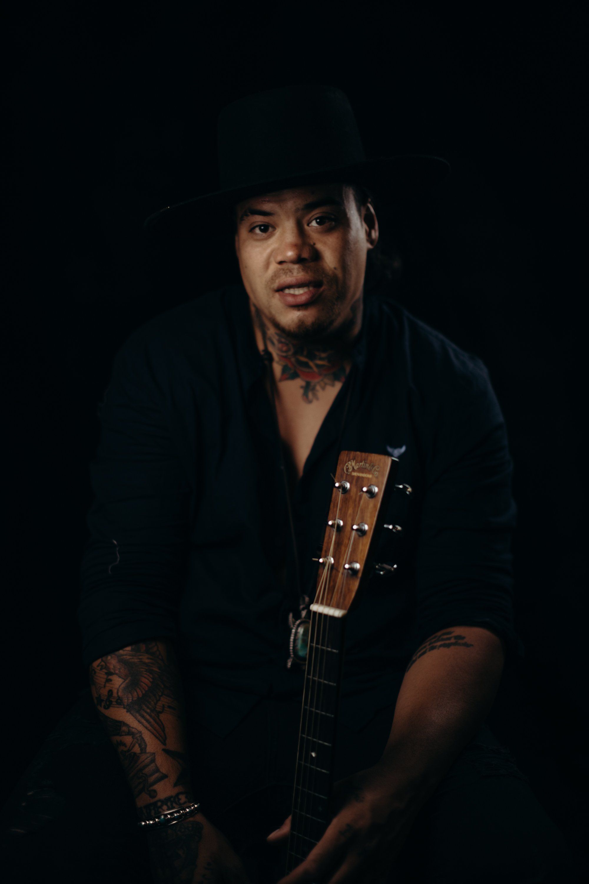 Man with tattooed arms holding acoustic guitar against dark background, wearing black hat and open shirt, dramatic studio lighting