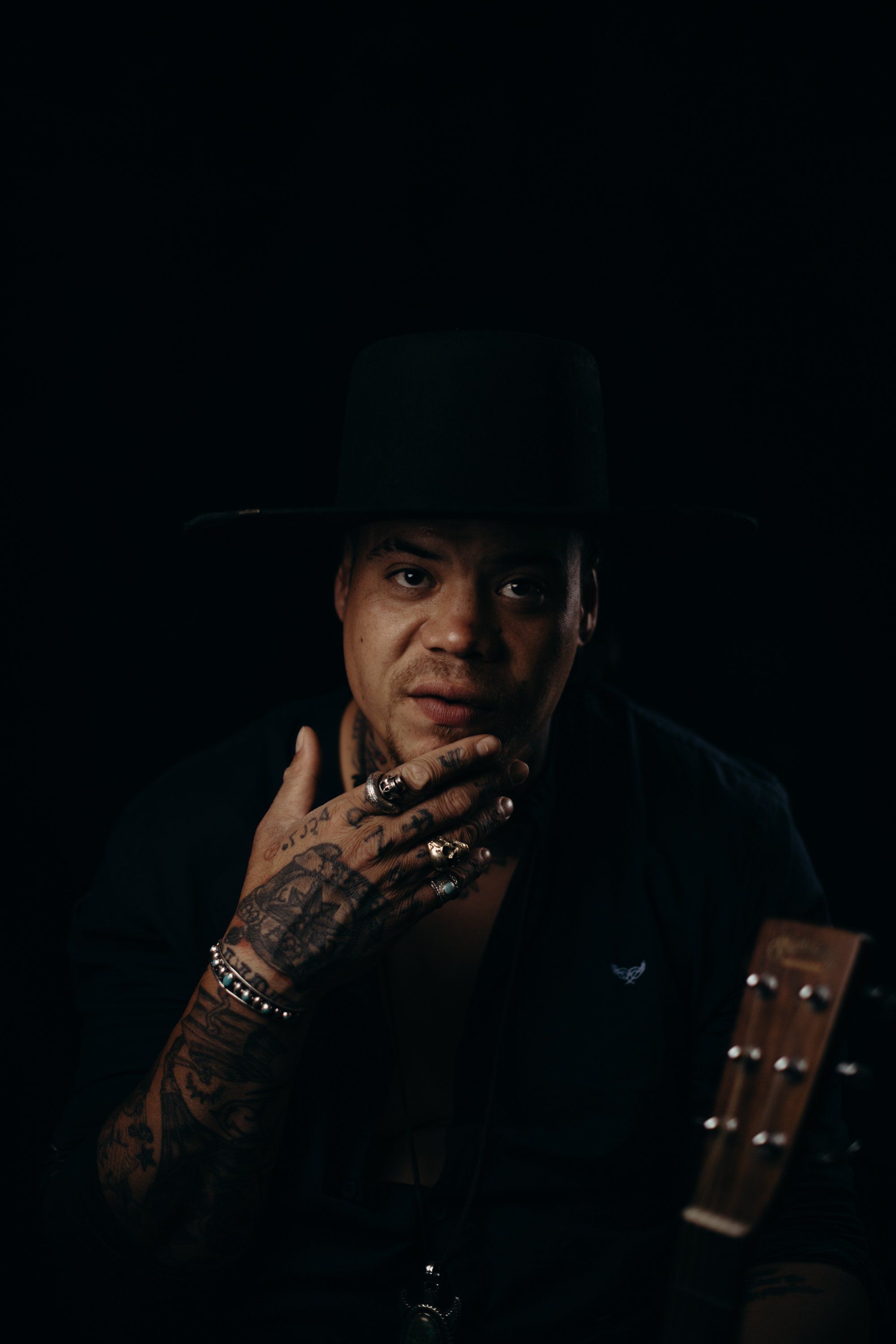 Man with tattooed hands and rings gazes thoughtfully against dark background, dramatic side lighting