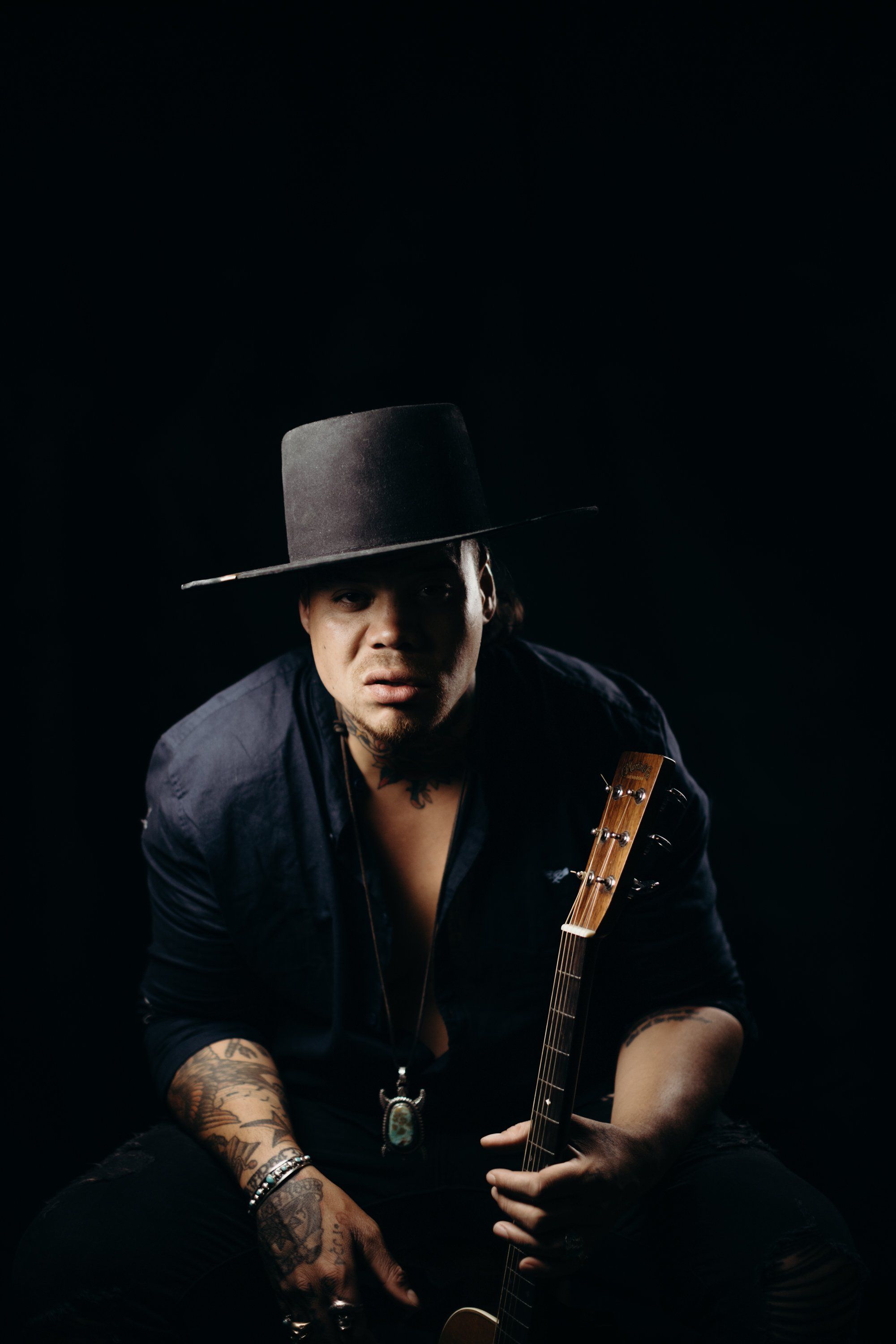 Man in black shirt and top hat holding acoustic guitar against dark background, dramatic side lighting