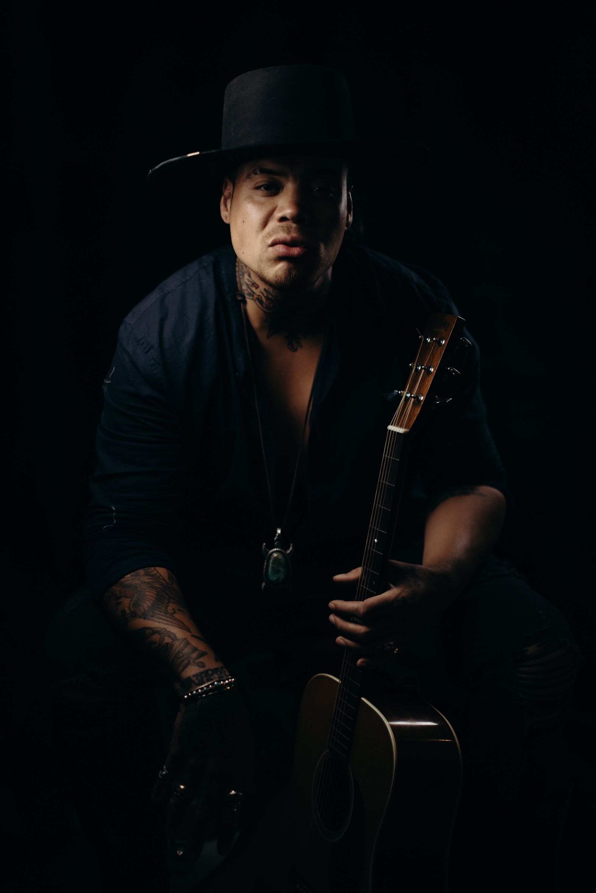 Man in black cowboy hat holding acoustic guitar against dark background, dramatic side lighting illuminating his face and tattooed arm