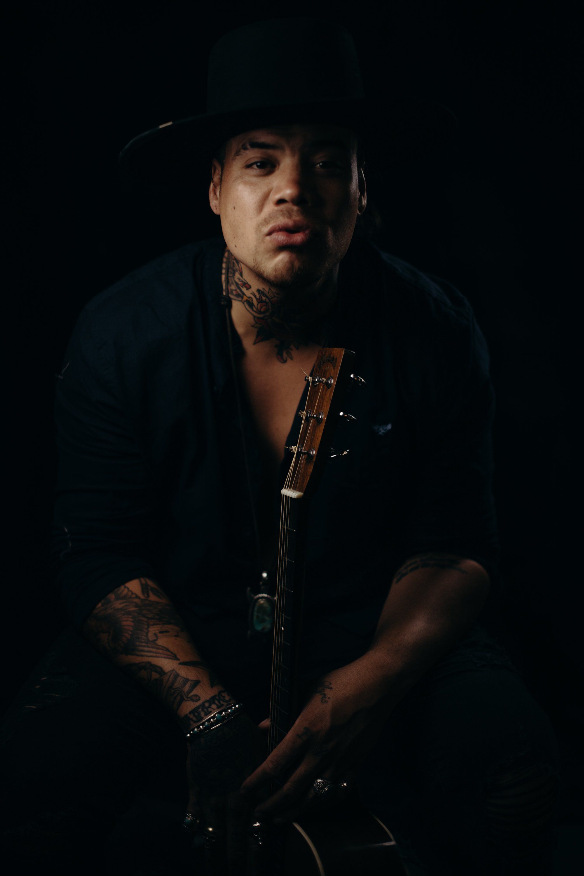 Tattooed man in dark clothing holding a guitar against black background, lit dramatically from the side