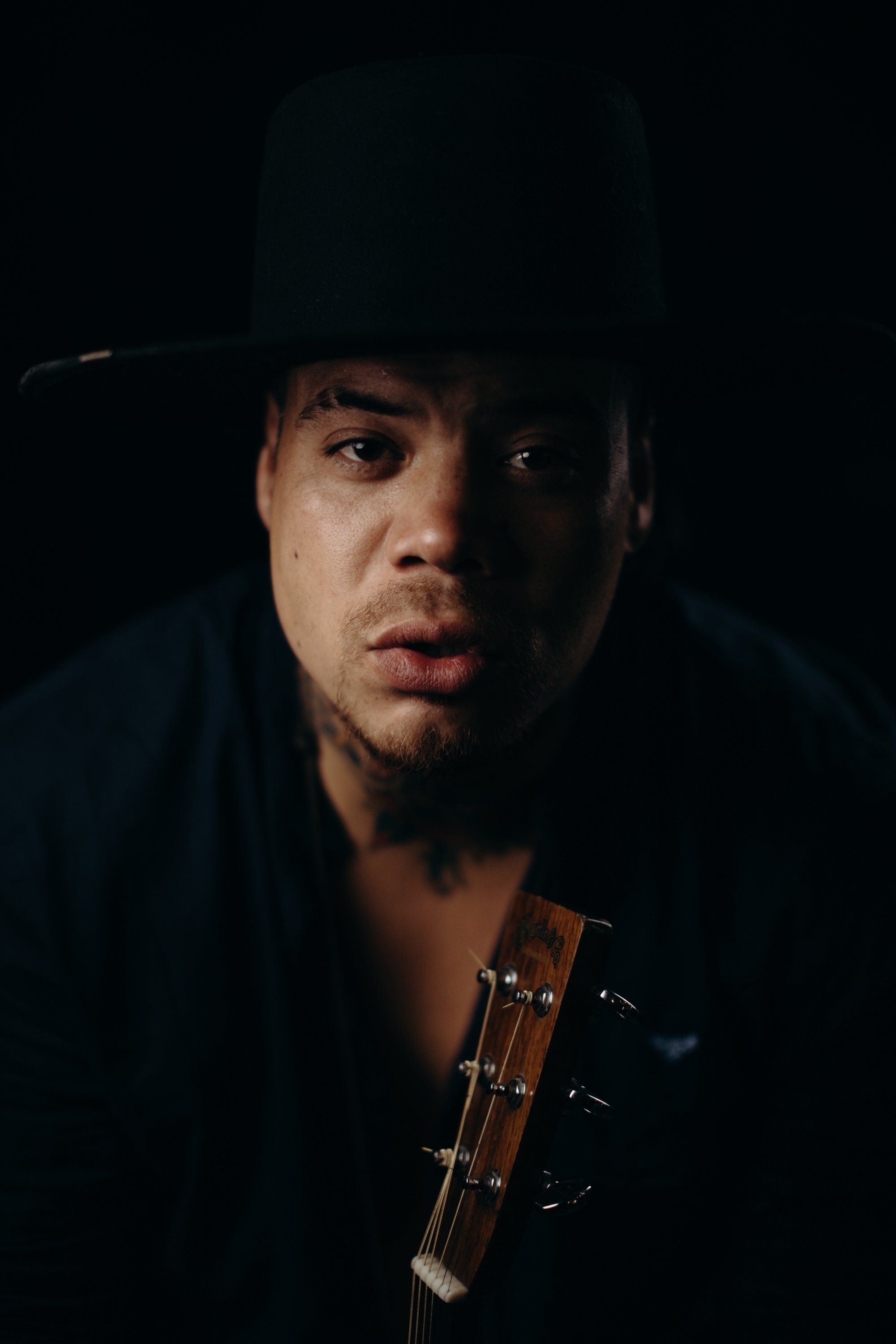 Man in dark hat and shirt holding guitar against black background, dramatically lit from one side