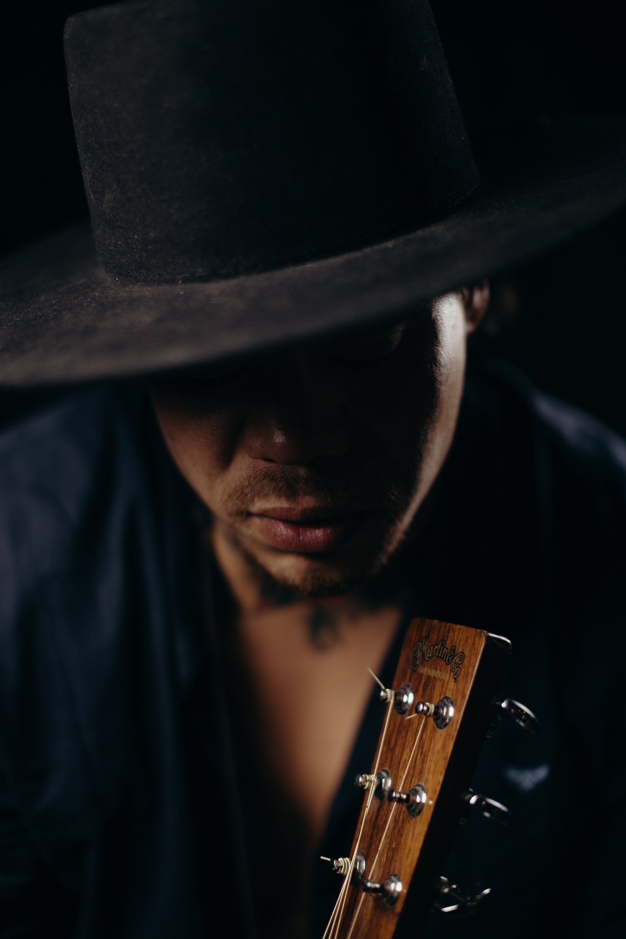 Man in black cowboy hat holding acoustic guitar, dramatically lit against dark background with warm light catching his face