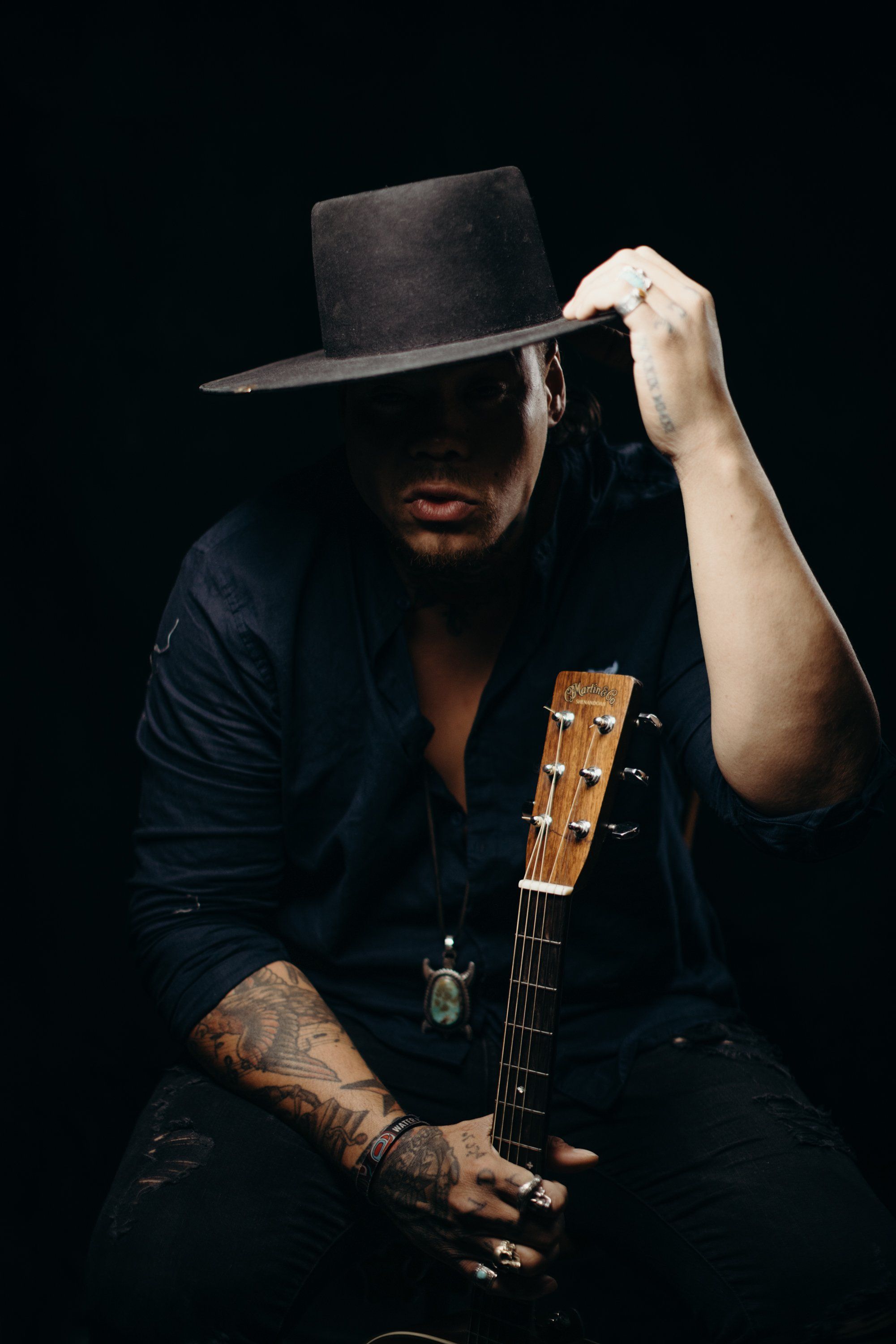 Bearded musician in black cowboy hat and shirt holds acoustic guitar against dark background, adjusting hat brim with contemplative expression