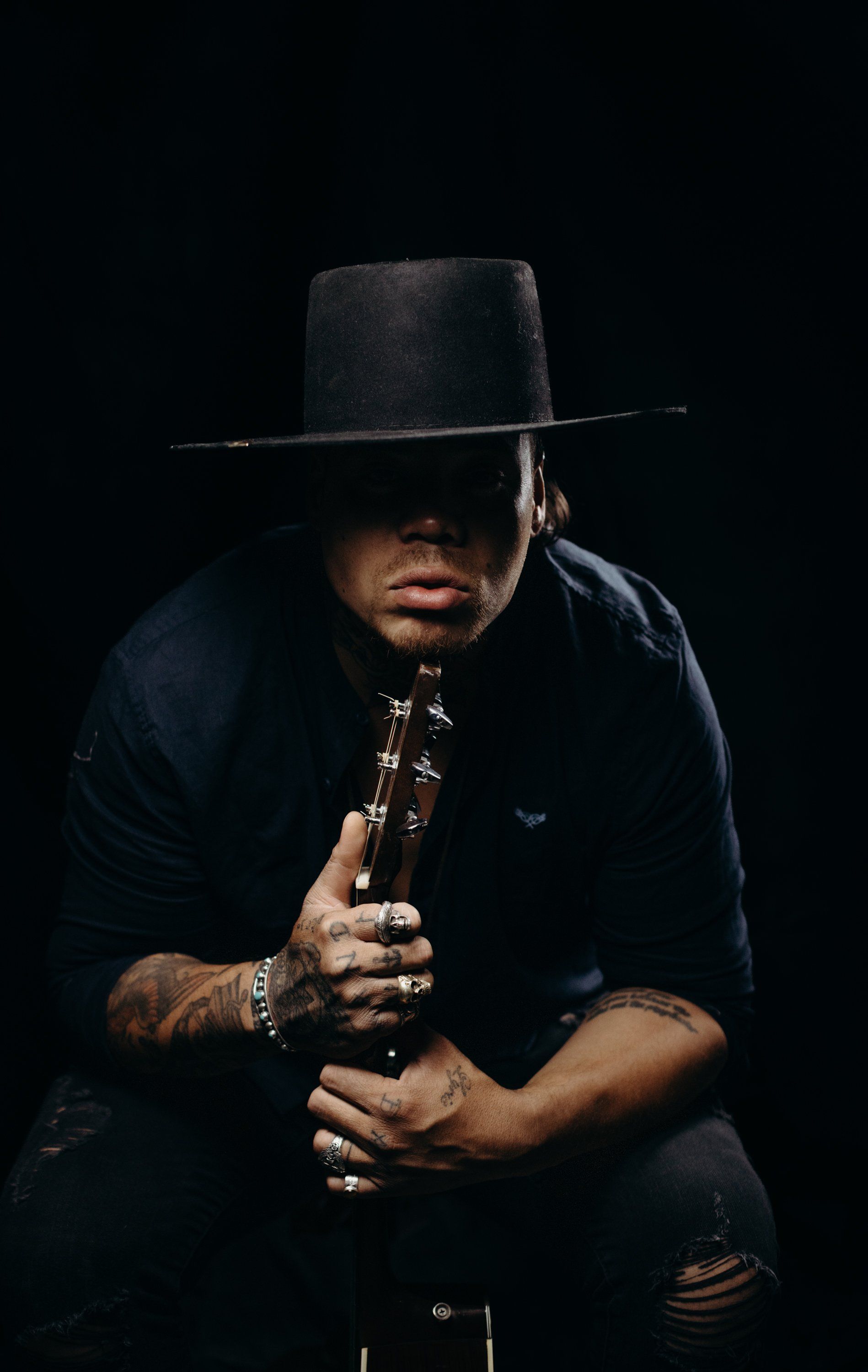 Man in black cowboy hat and shirt holds ornate cross pendant against dark background, dramatic side lighting