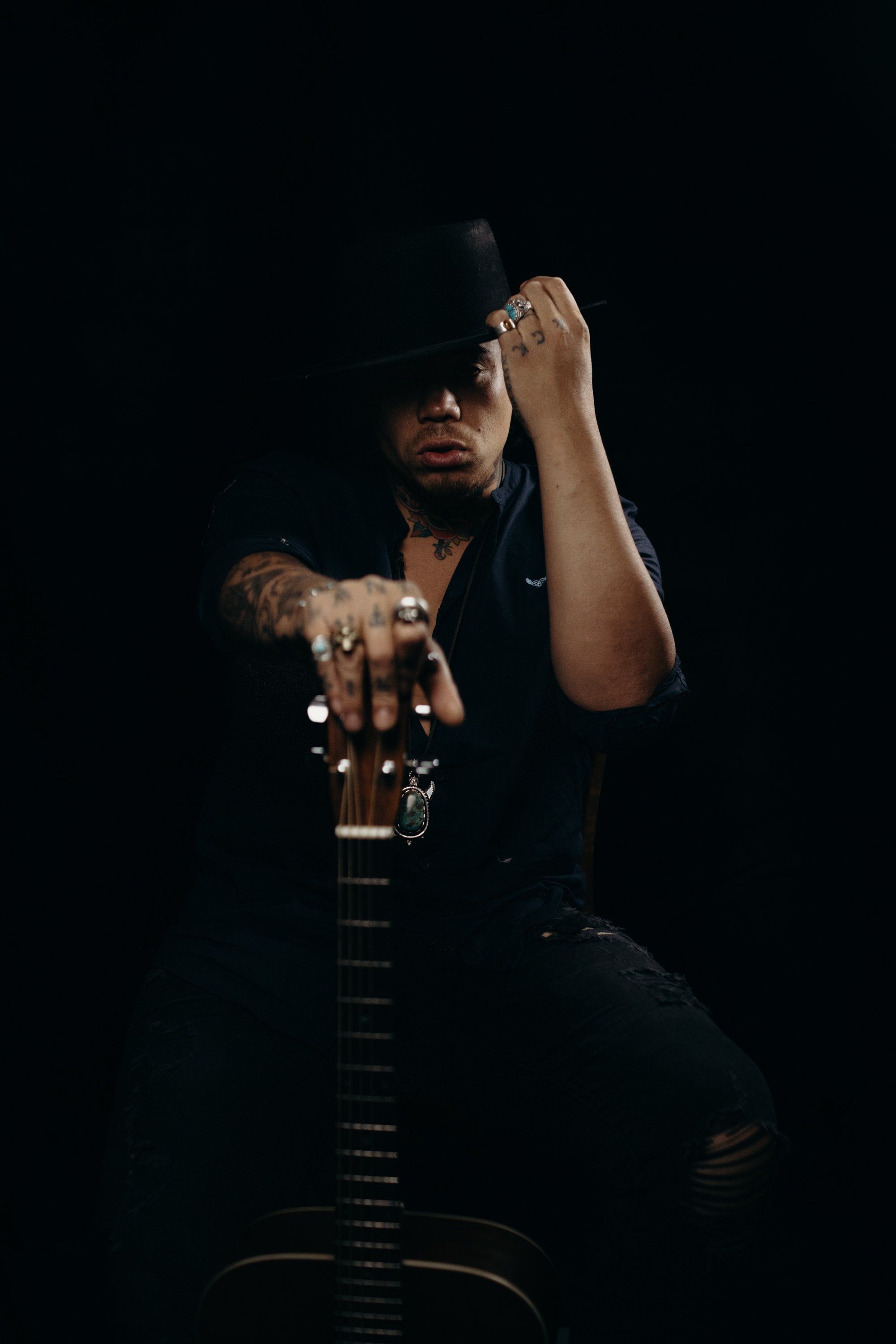 Tattooed musician holding electric guitar against black background, adjusting wide-brimmed hat in dramatic studio lighting