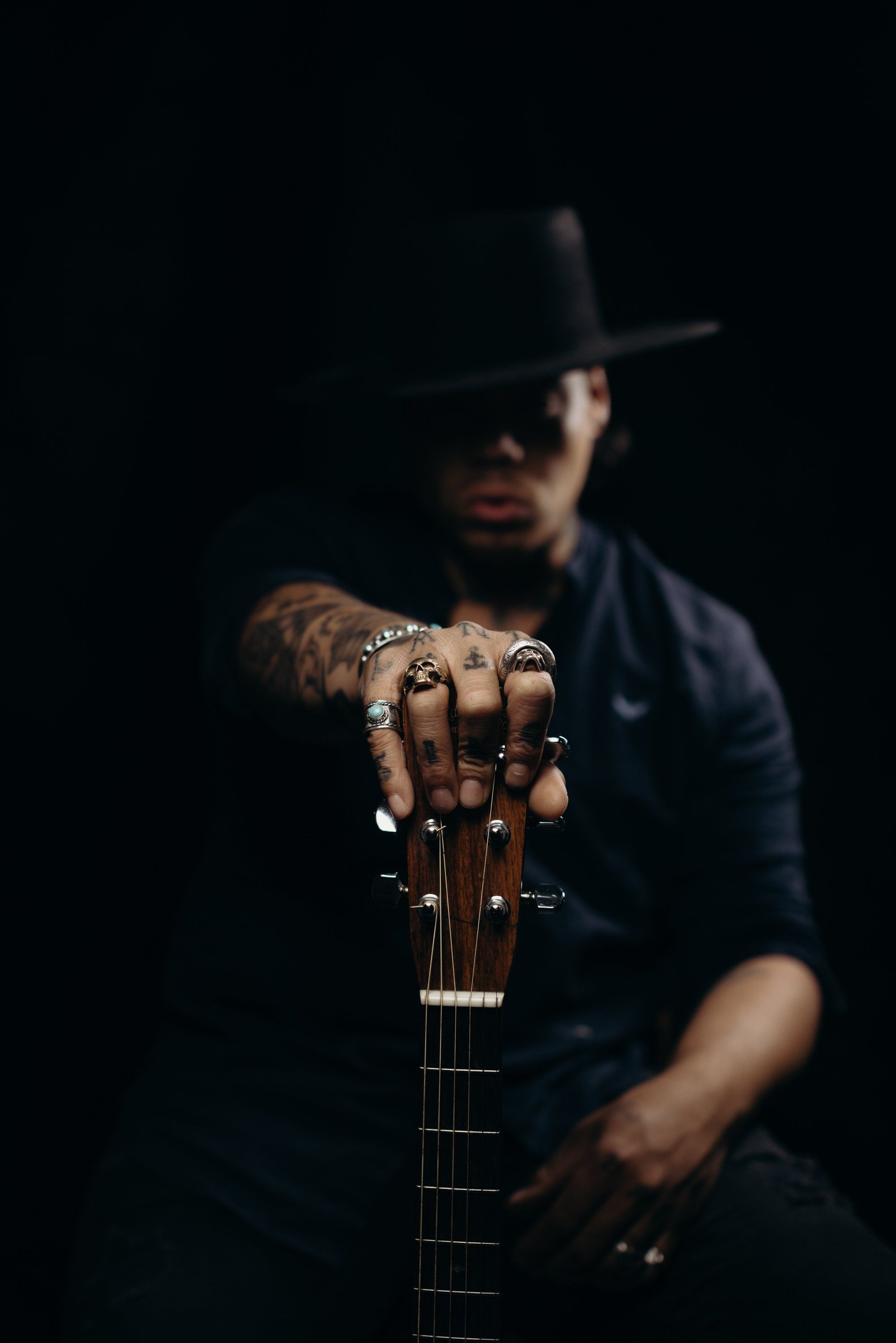 Man in dark clothing and wide-brimmed hat extends acoustic guitar toward camera against black background, tattoos visible on hands