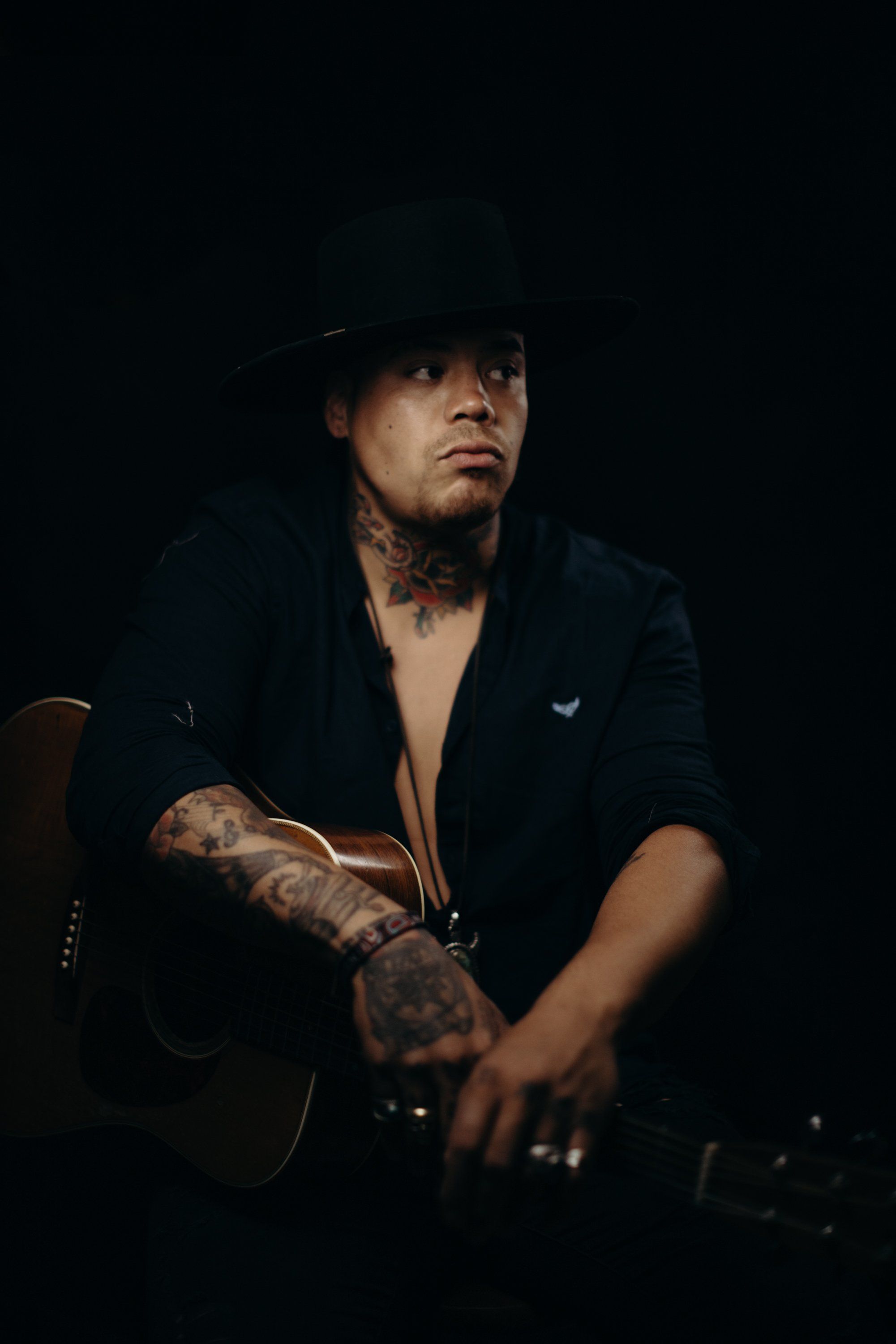 Tattooed man in dark blazer and hat against black background, dramatic side lighting casting shadows across his face and arms