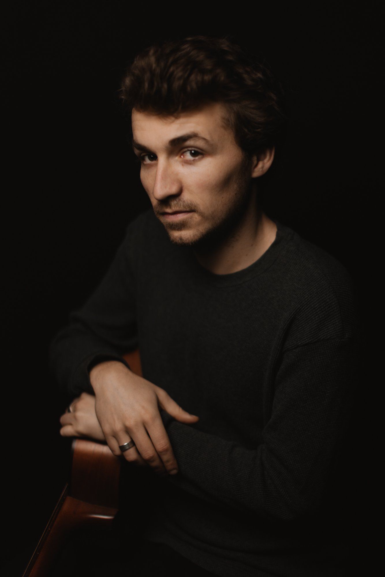 Man in black long-sleeve shirt against dark background, posed with arms crossed, warm studio lighting illuminating his face and hand