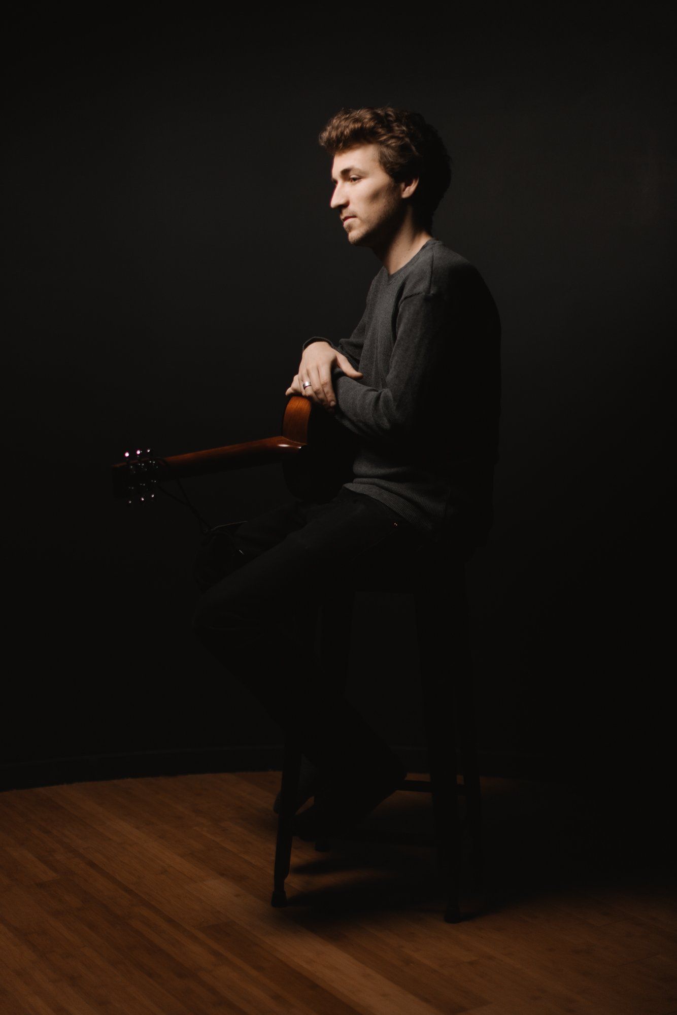 Man in dark clothing sitting with violin against black backdrop, warm wooden floor visible below