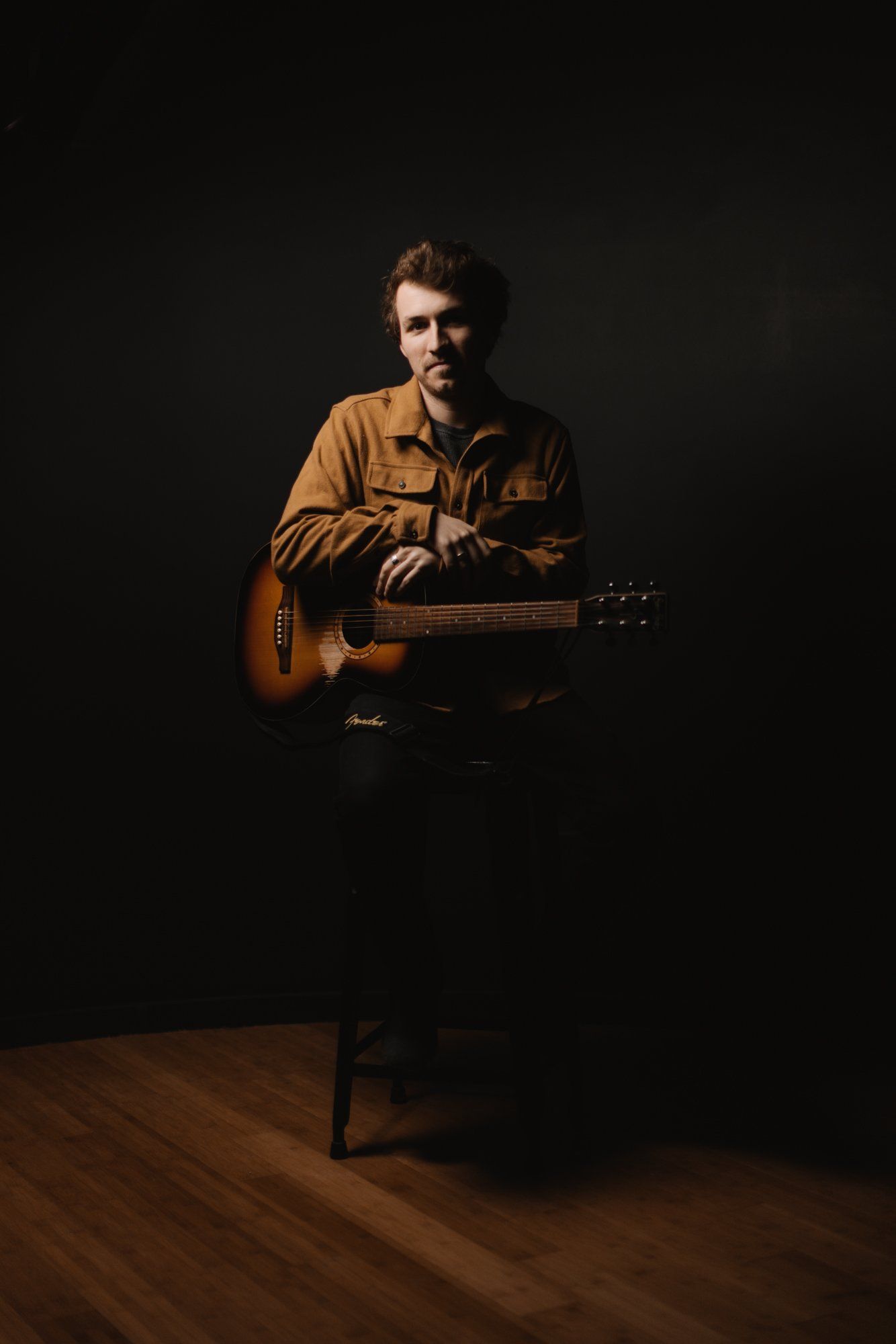 Musician with acoustic guitar in warm light against dark background, seated in contemplative pose