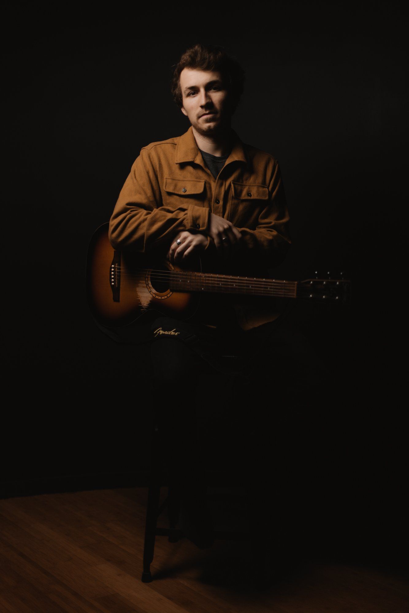 Musician in warm-toned shirt holds acoustic guitar against dark background, dramatic side lighting creating intimate studio portrait