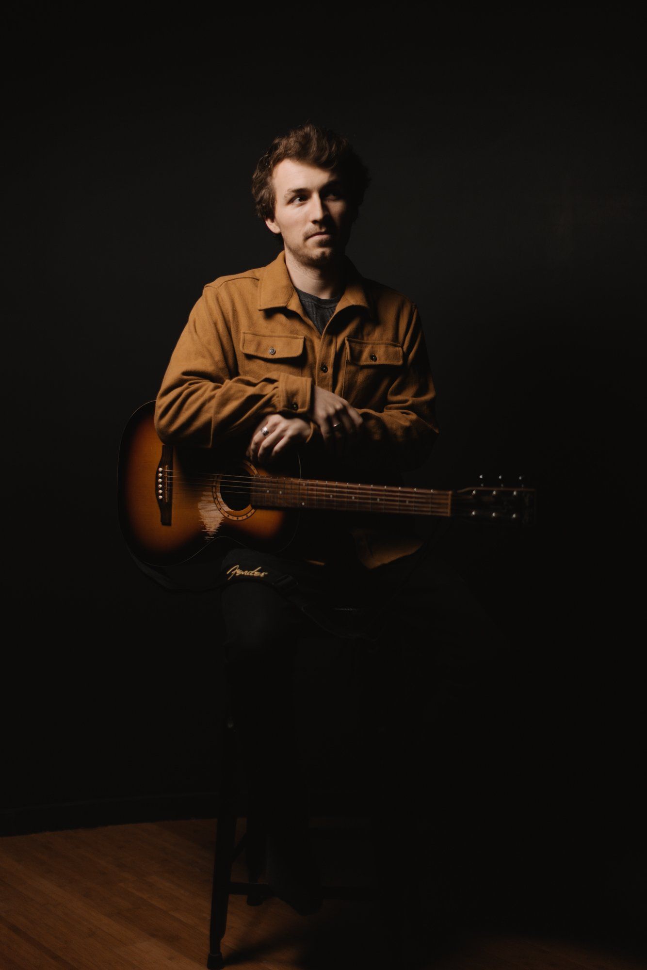 Man in mustard shirt holding acoustic guitar against dark background, warm studio lighting