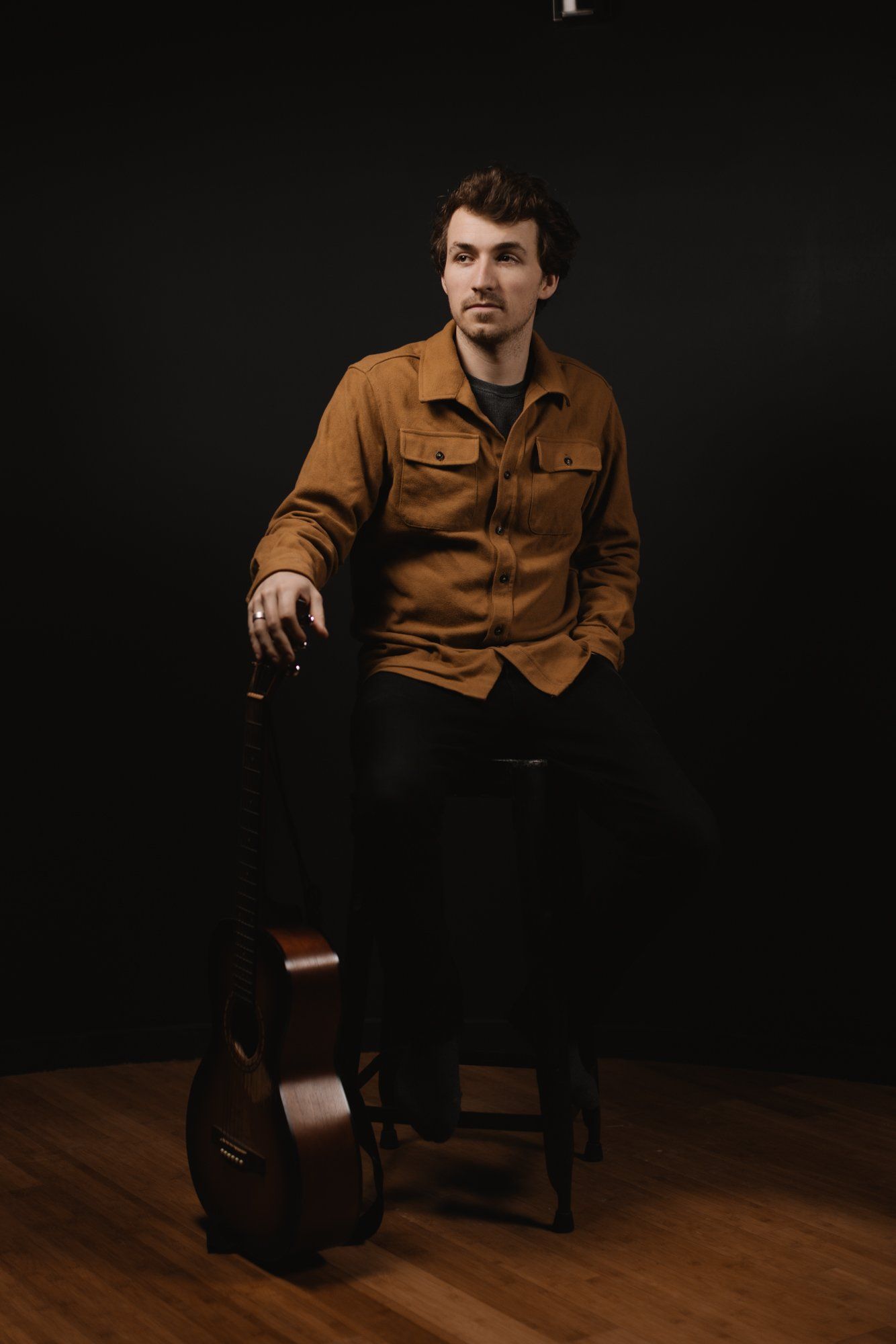 Man in mustard shirt and black ripped jeans sits with acoustic guitar against dark backdrop on wooden floor