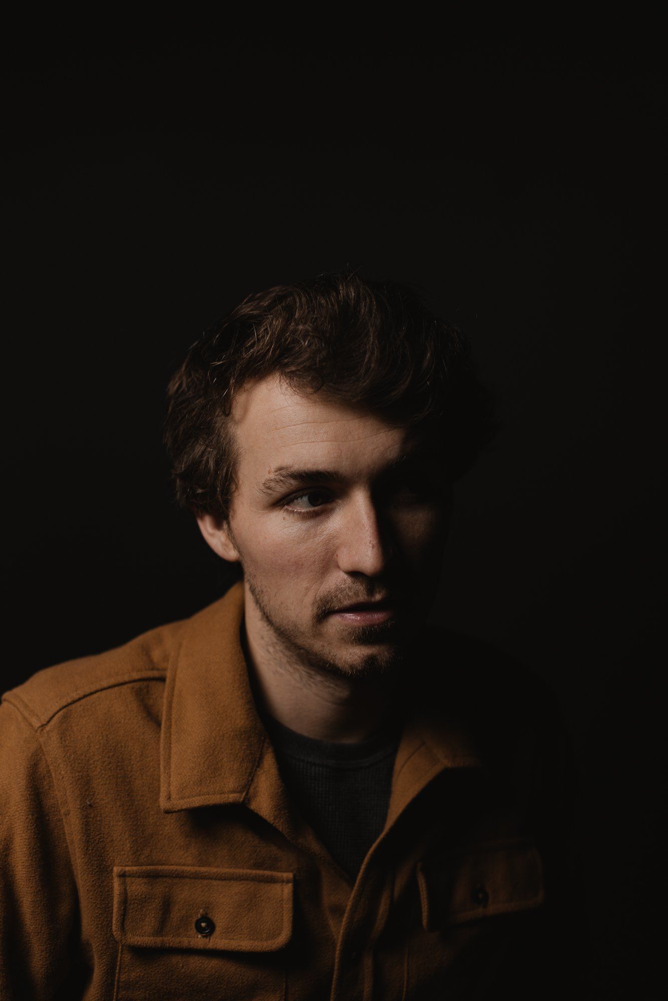 Man in caramel jacket against dark background, side-lit portrait with warm golden light catching his contemplative gaze