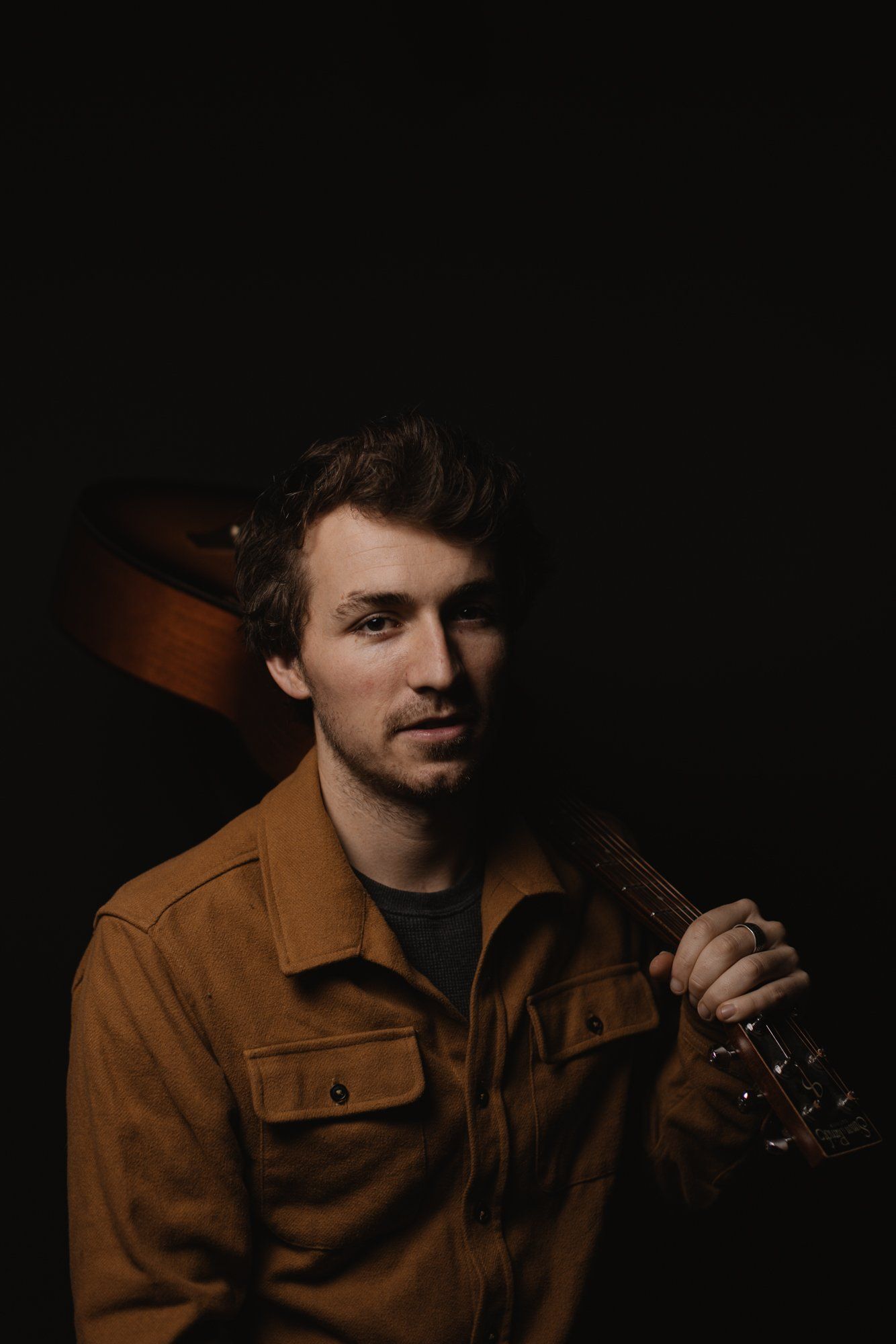Man in tan work shirt holding guitar against dark background, warm studio lighting casting shadows
