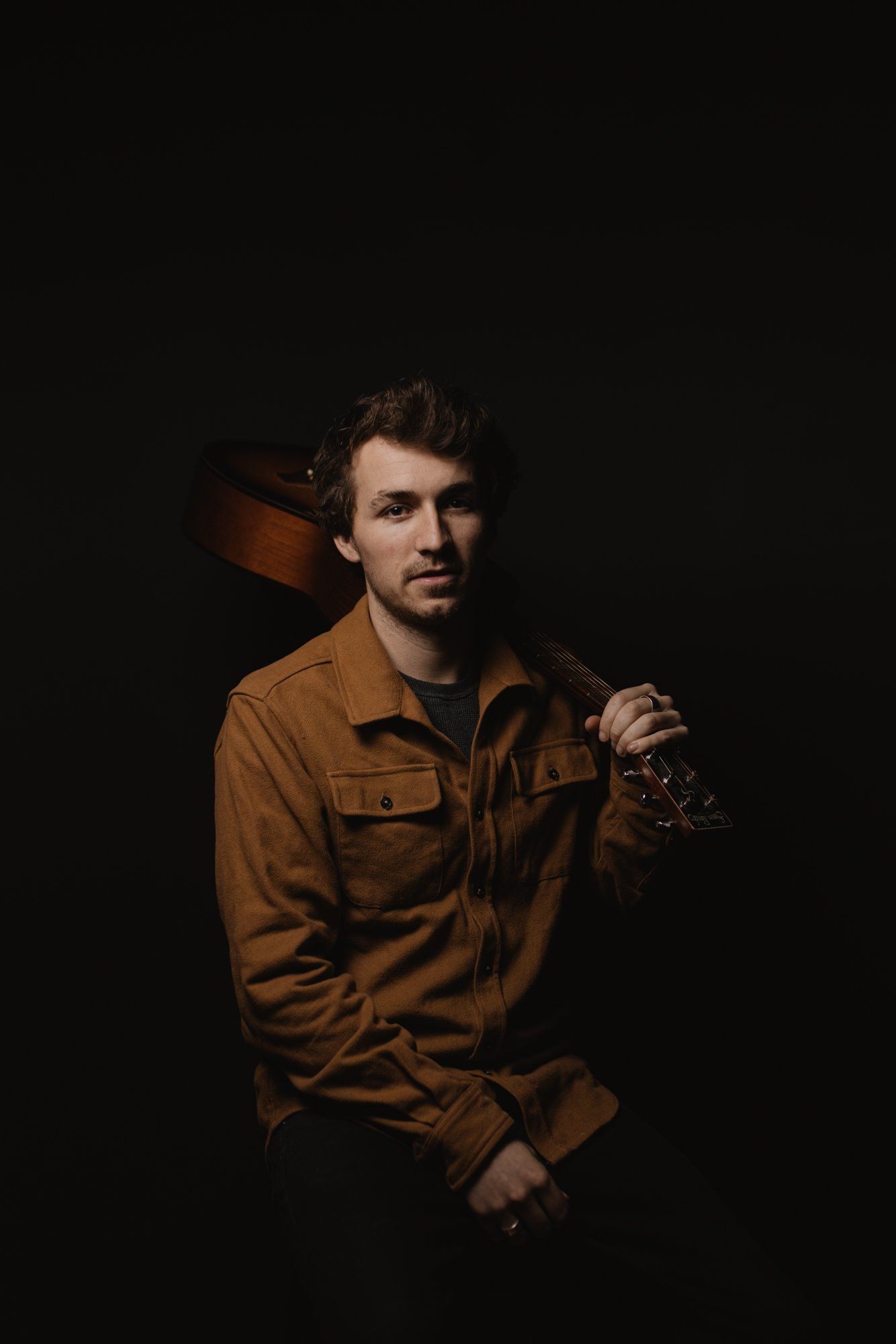 Man in rust-colored shirt with guitar, dramatically lit against black background, gazing thoughtfully to side