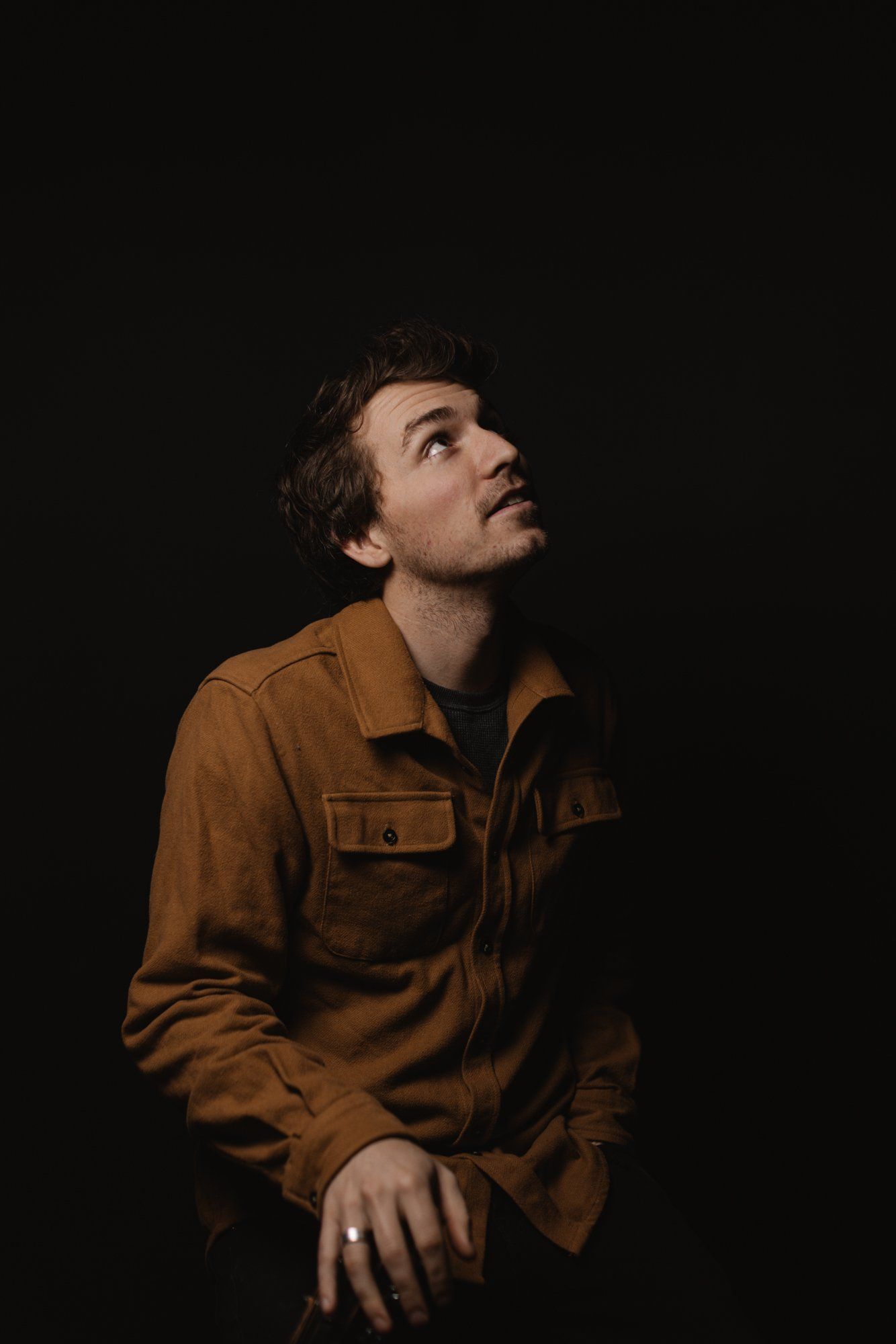 Man in burnt-orange shirt gazes upward against black background, warm studio lighting highlighting contemplative profile
