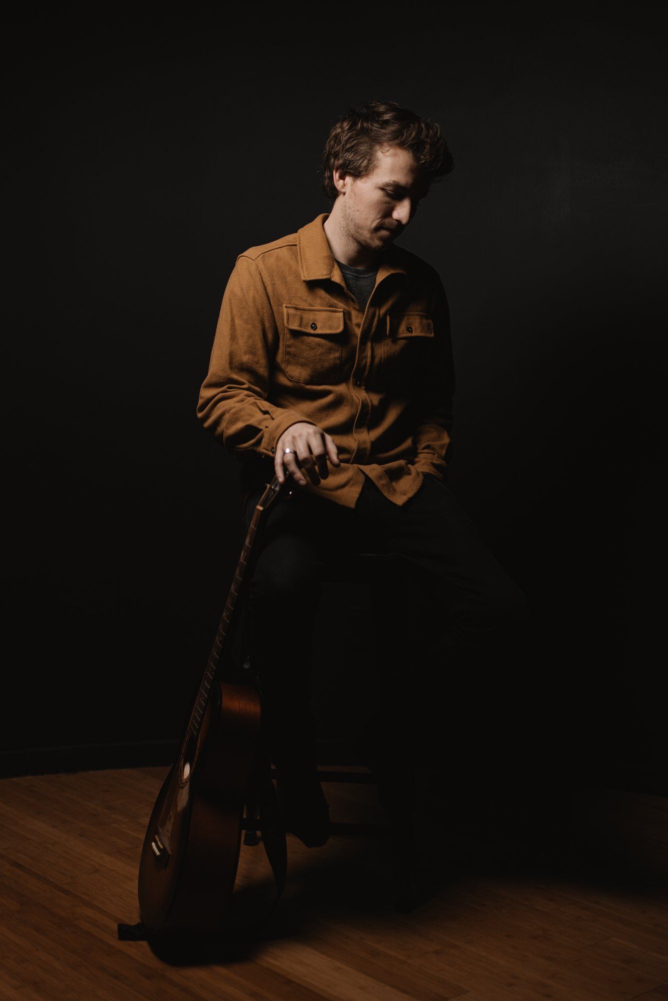 Man in tan shirt holding guitar, dramatically lit against black background on wooden floor