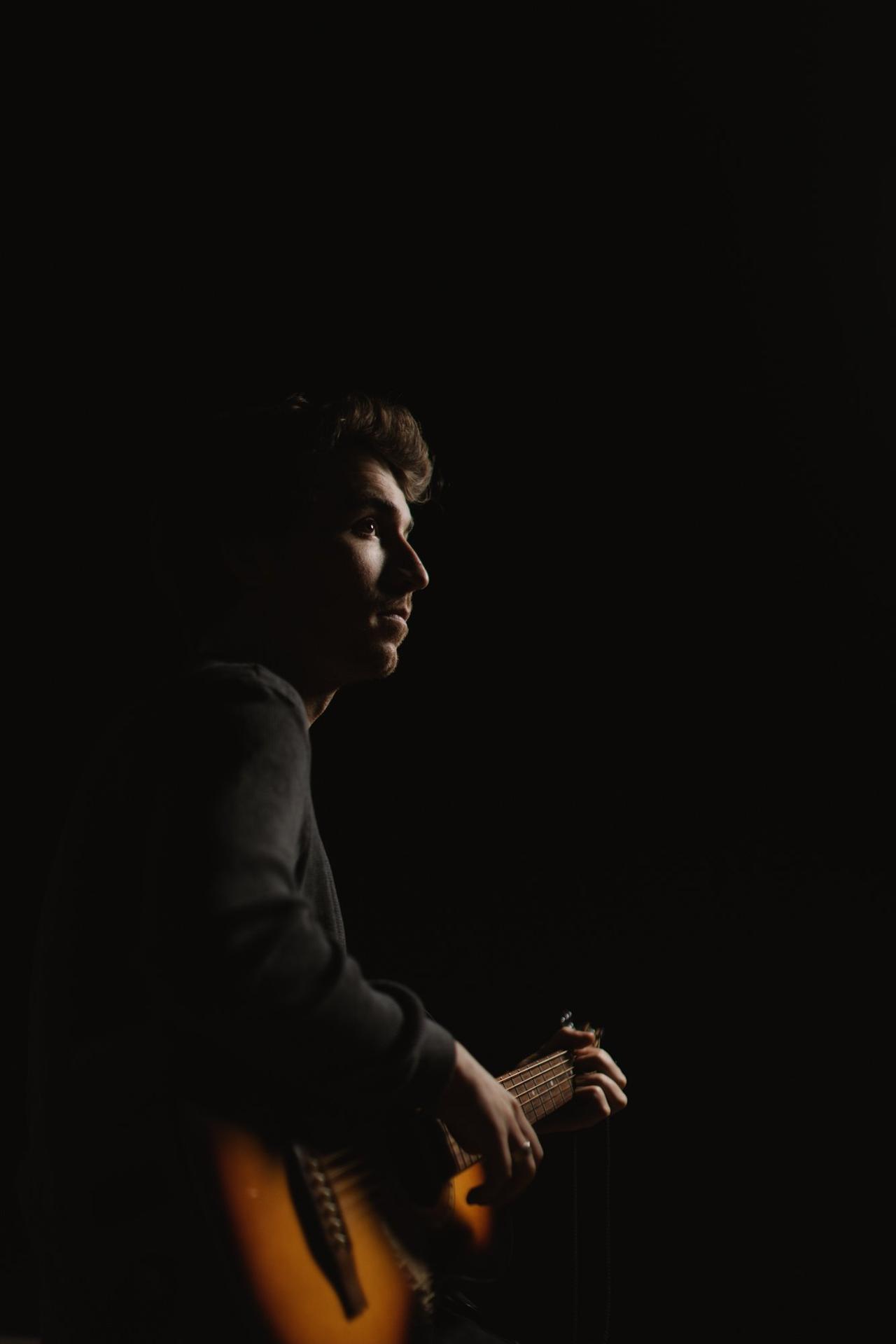 Musician in profile against black background, hands on acoustic guitar, dramatic side lighting on face