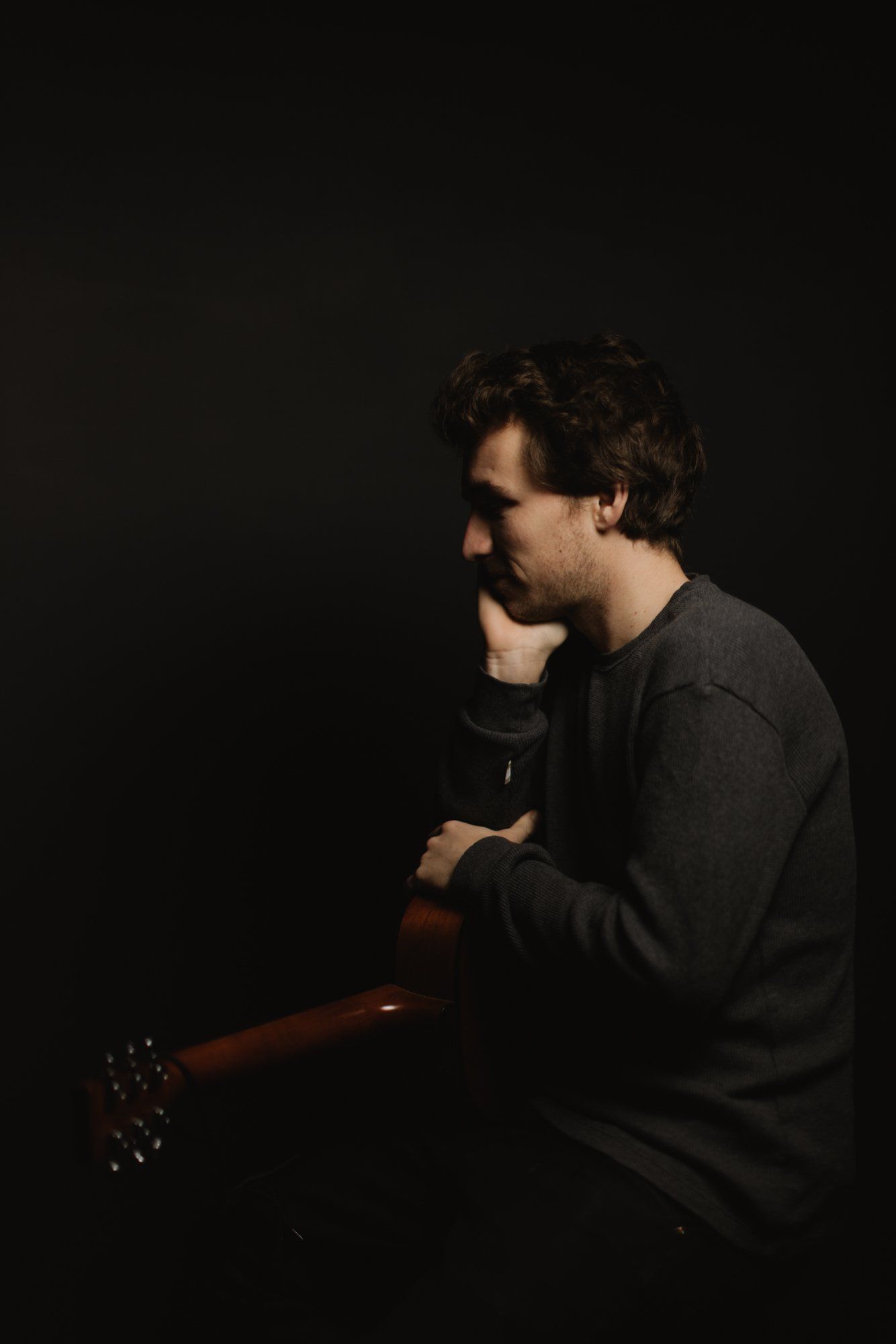 Man in black shirt holding acoustic guitar against dark background, side profile with warm studio lighting