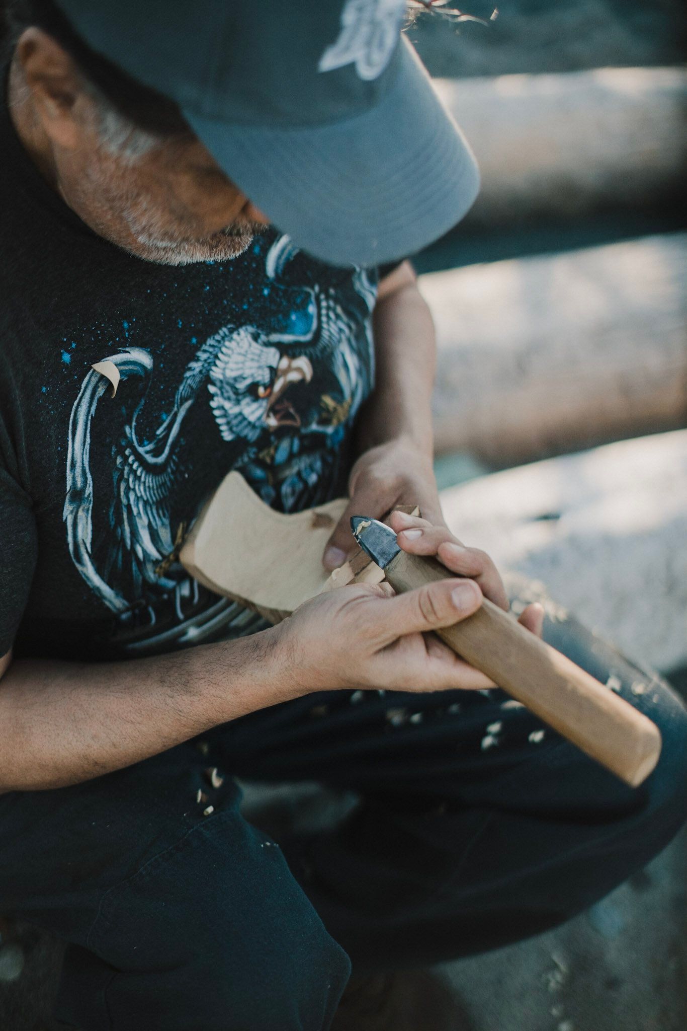 Hands carving wood with a chisel, wearing a blue cap and dark graphic tee, sawdust caught in golden afternoon light