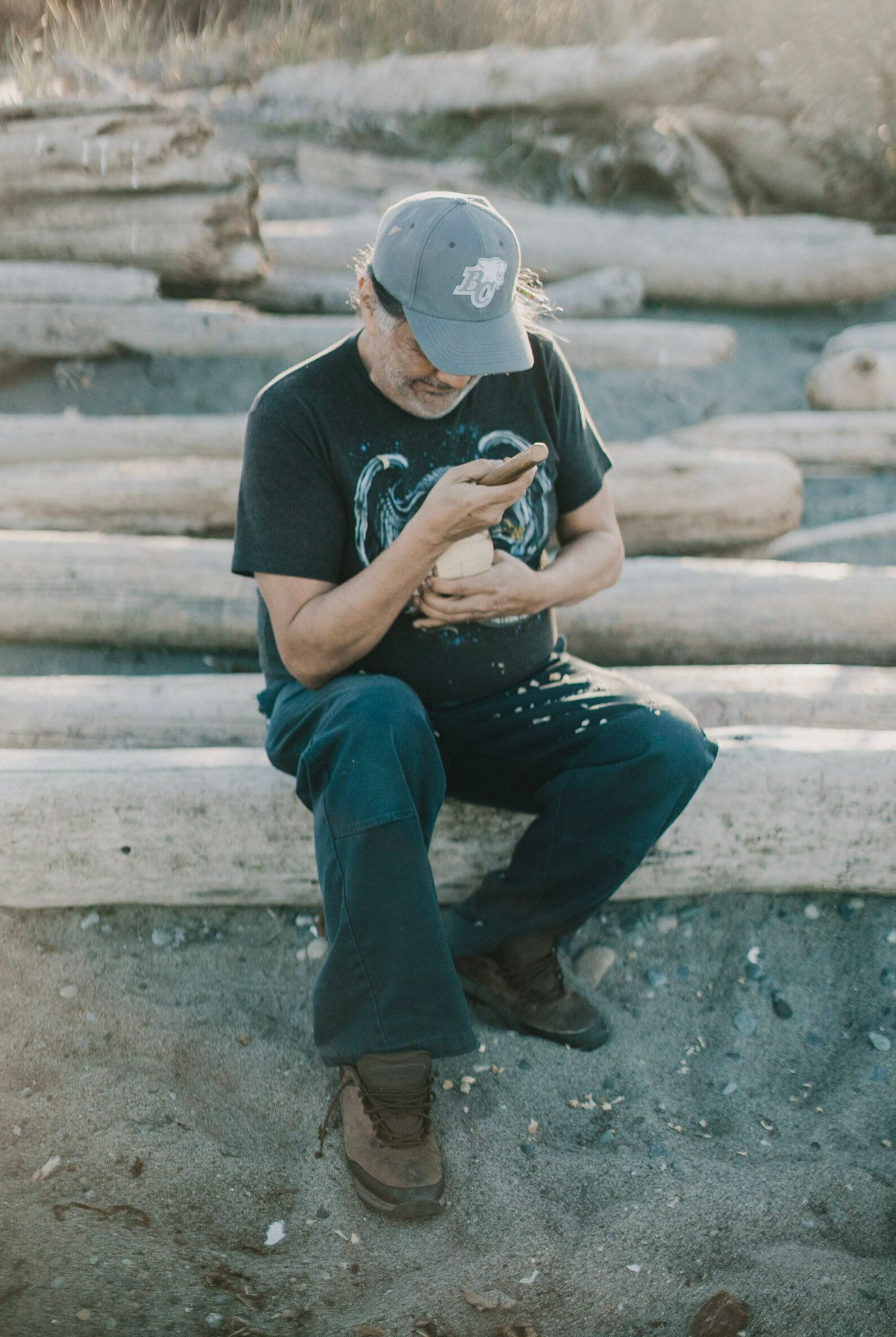 Person in cap and dark shirt crouches on driftwood, examining something in their hands amid weathered logs and beach sand