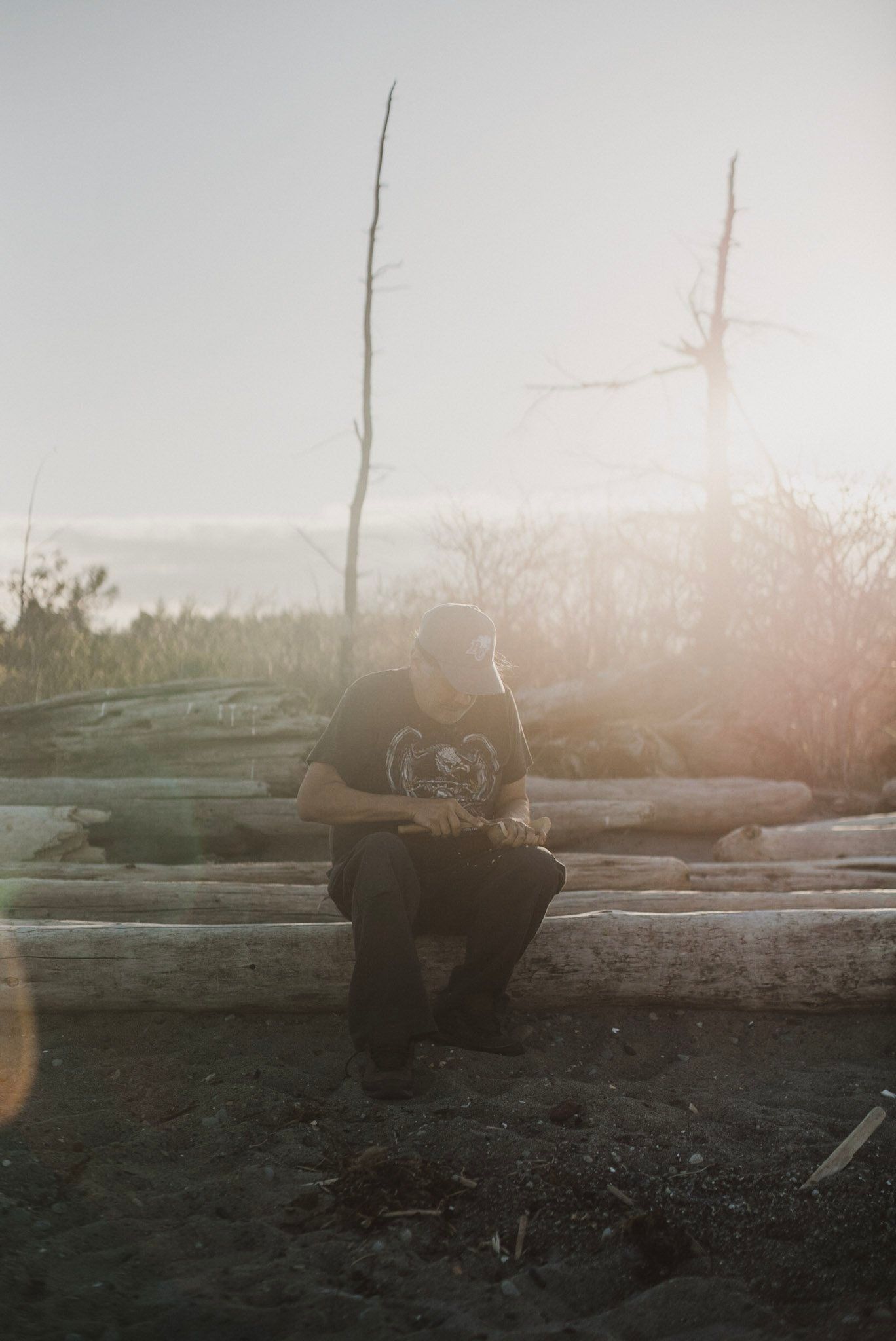 Solitary figure seated on weathered driftwood in misty morning light, surrounded by bare branches and soft fog
