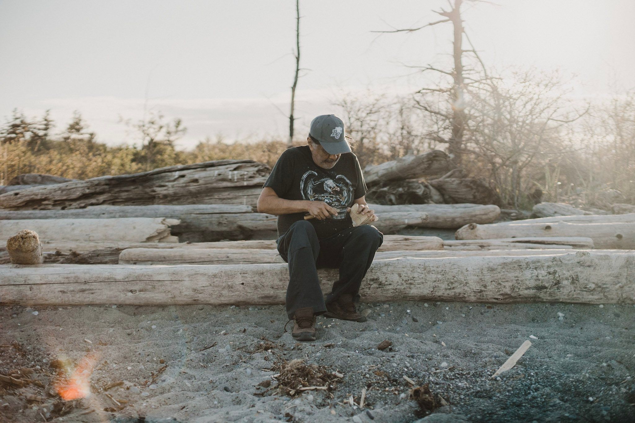 Man in dark shirt crouched by small campfire among driftwood logs on misty beach