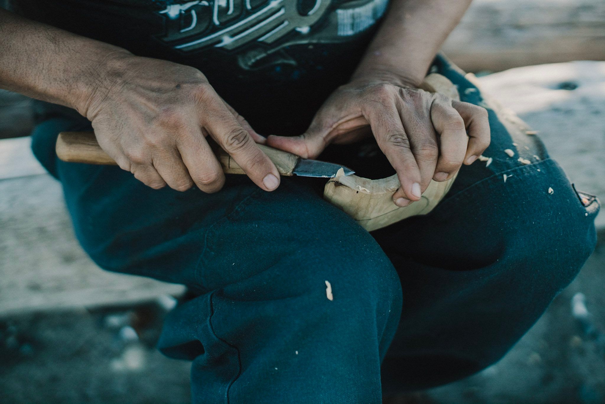 Weathered hands carving wood with a knife, seated outdoors in natural light with wood shavings visible