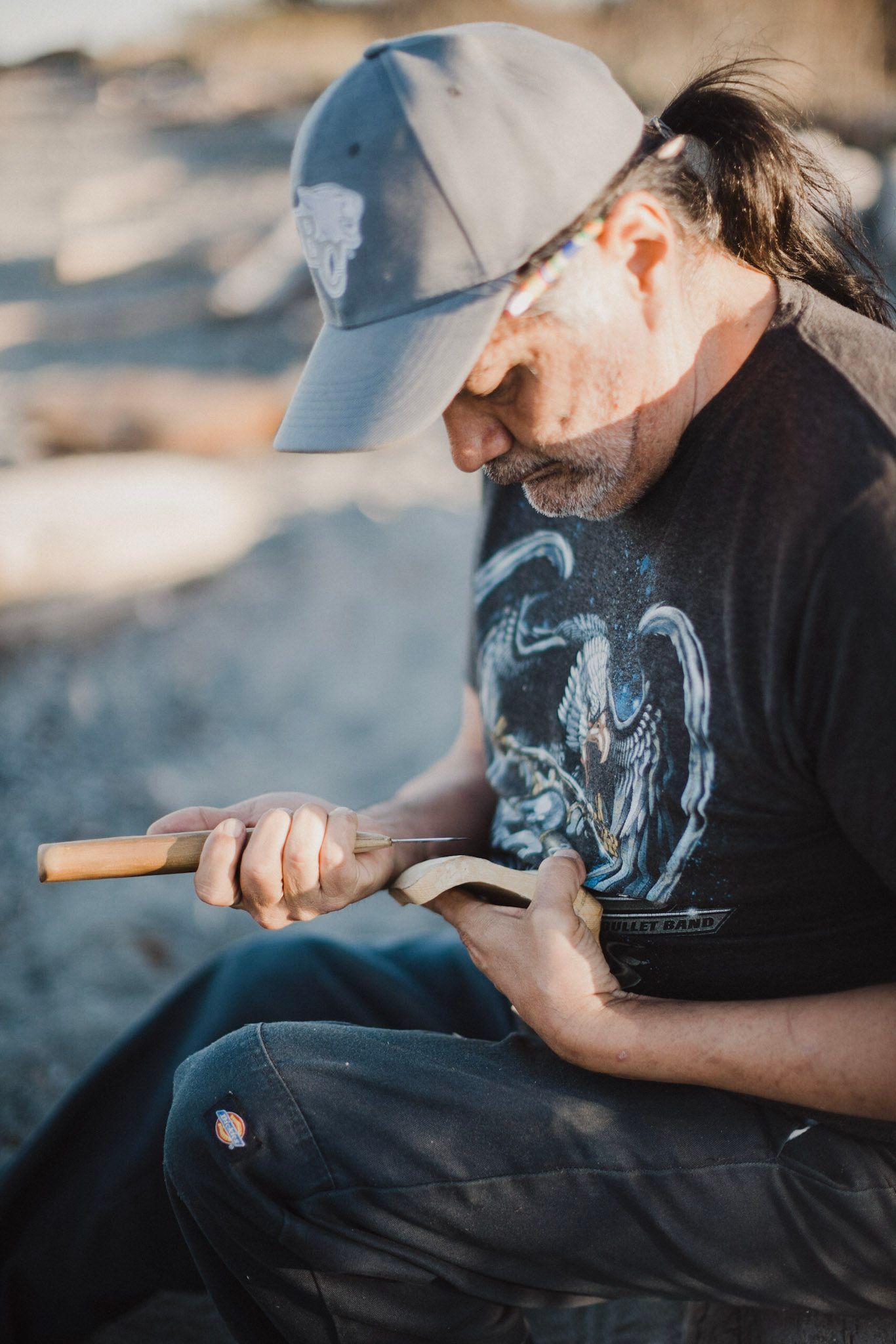 Bearded man in cap and graphic tee holds carving tools while seated outdoors in dappled sunlight, focused on his craft.