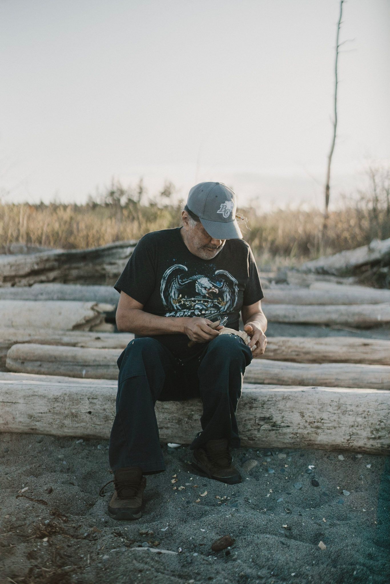 Man in black band tee and cap sitting on driftwood by misty water, evergreen forest beyond