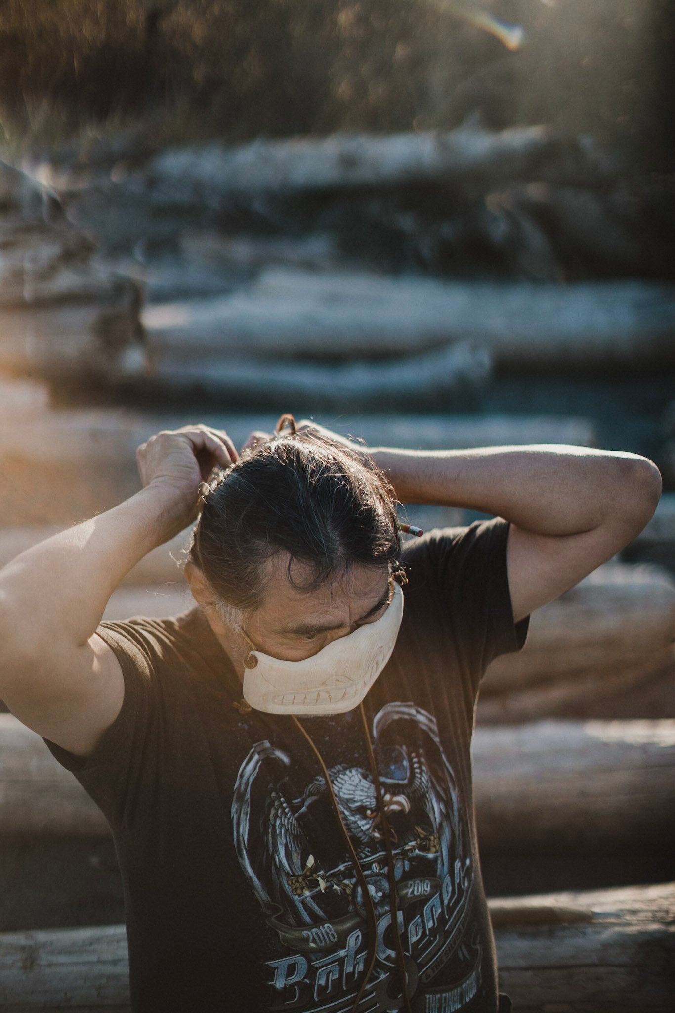 Young man in graphic tee with hands behind head, golden hour light on weathered logs, contemplative expression