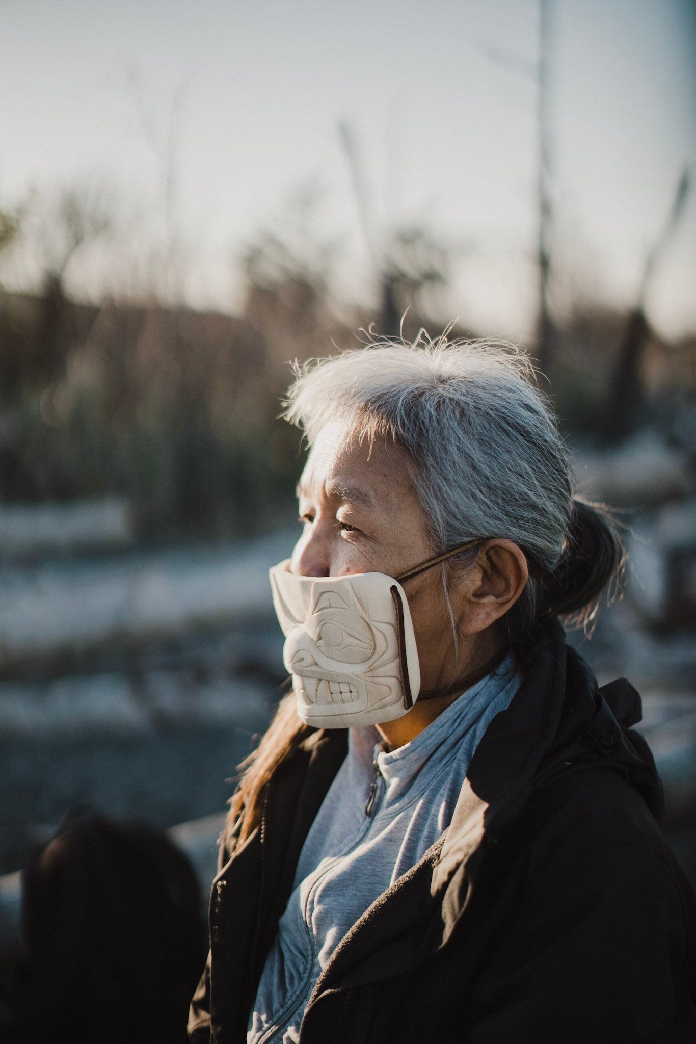 Elderly woman in white mask and black jacket gazes thoughtfully into golden sunlight, bare trees softly blurred behind her