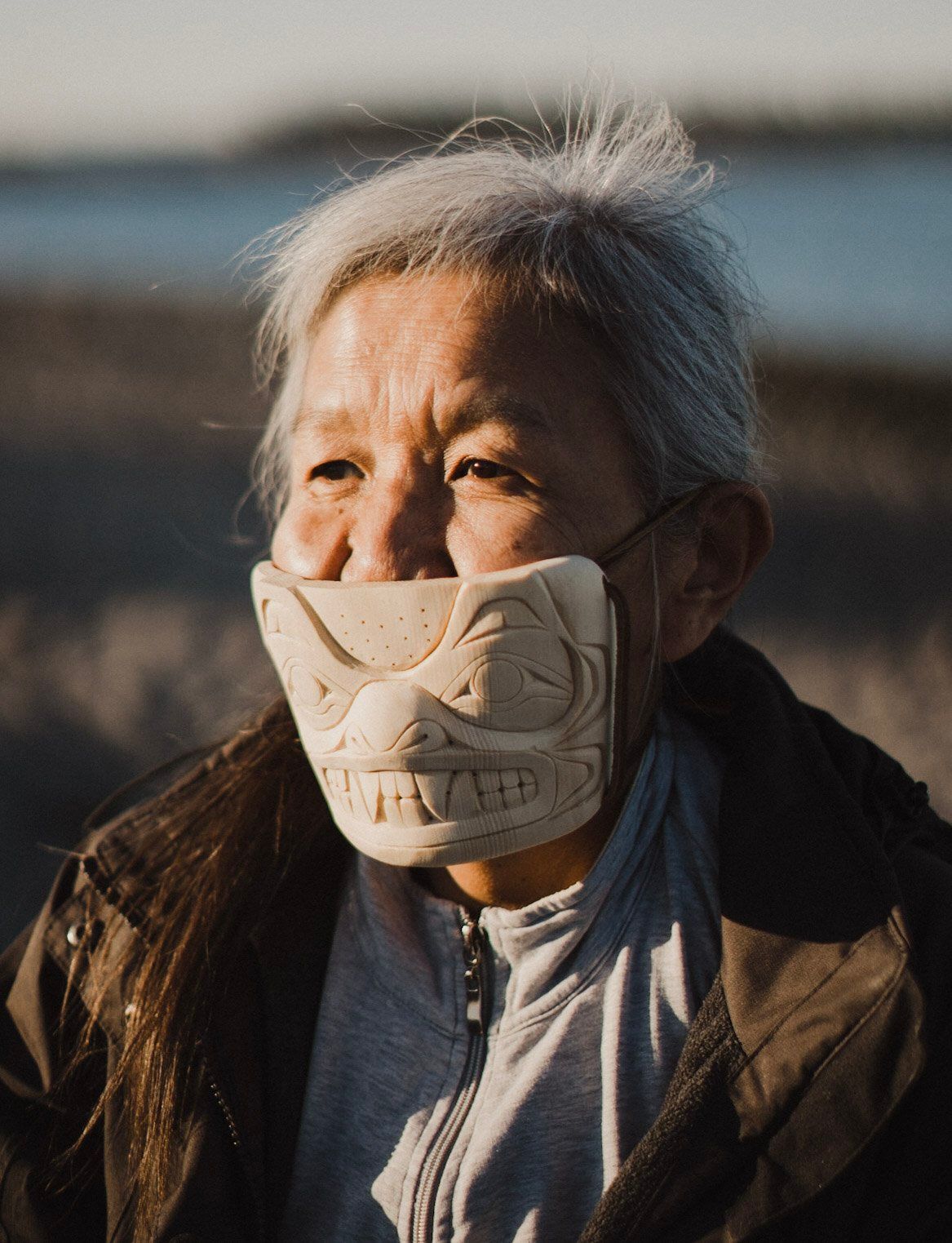 Elder wearing carved wooden mask with formline design, backlit by golden hour sun with blurred waterfront behind