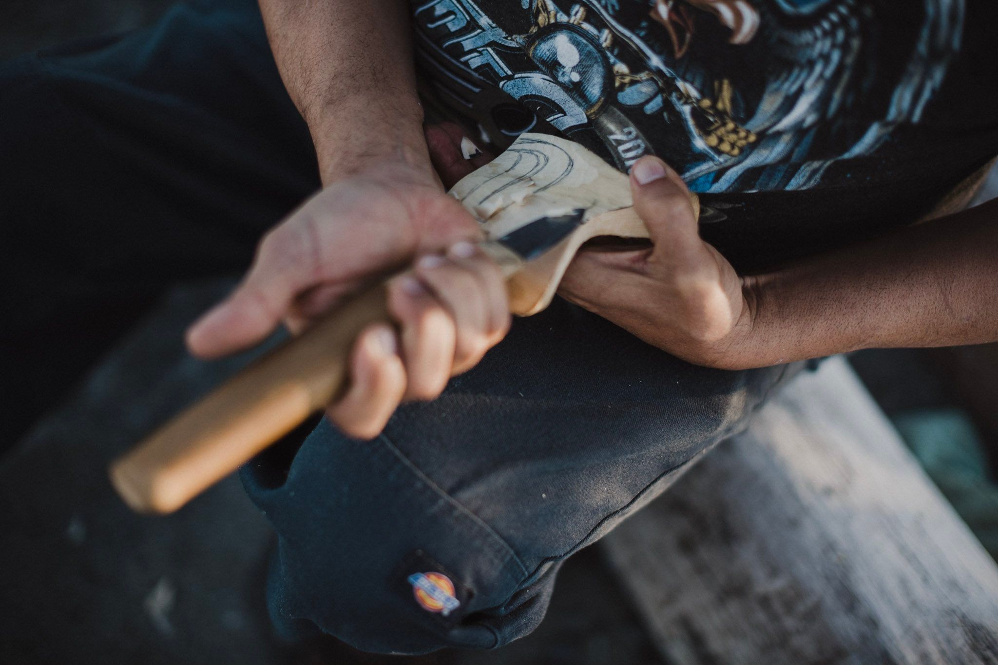 Carver's weathered hands grip a wooden tool against denim, golden sunlight illuminating the craftsmanship and worn details of his work