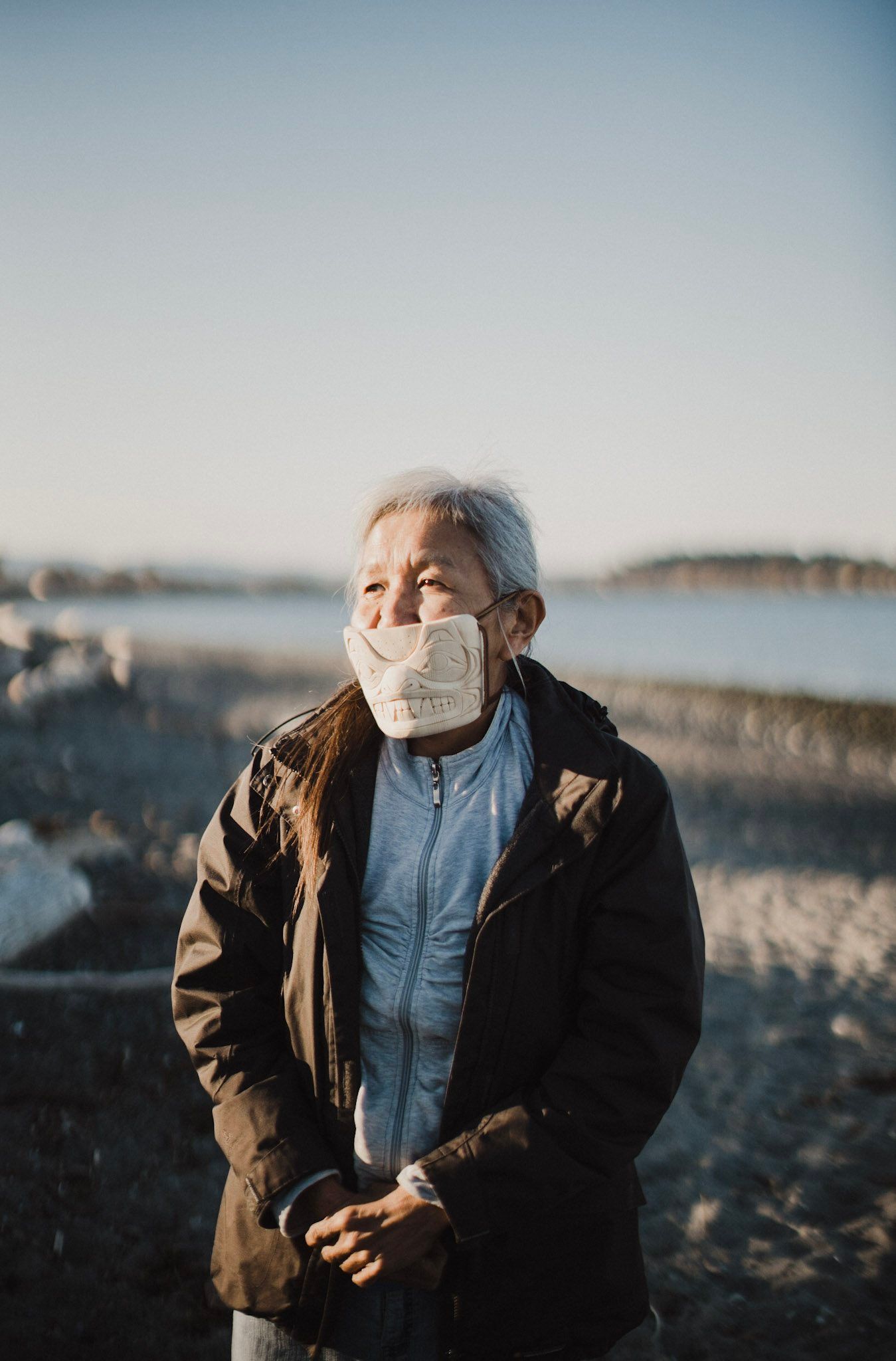 Elderly man wearing a mask and brown jacket stands on a rocky beach with calm water and forested shoreline behind him under clear sky