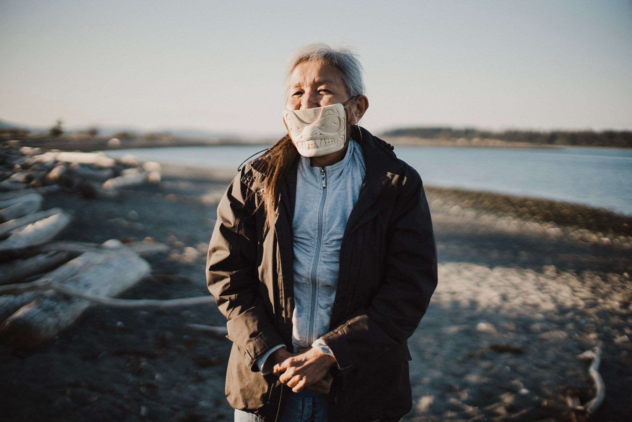 Elder with gray hair wearing a cloth mask and dark jacket stands on a rocky beach with calm water and distant shoreline behind them