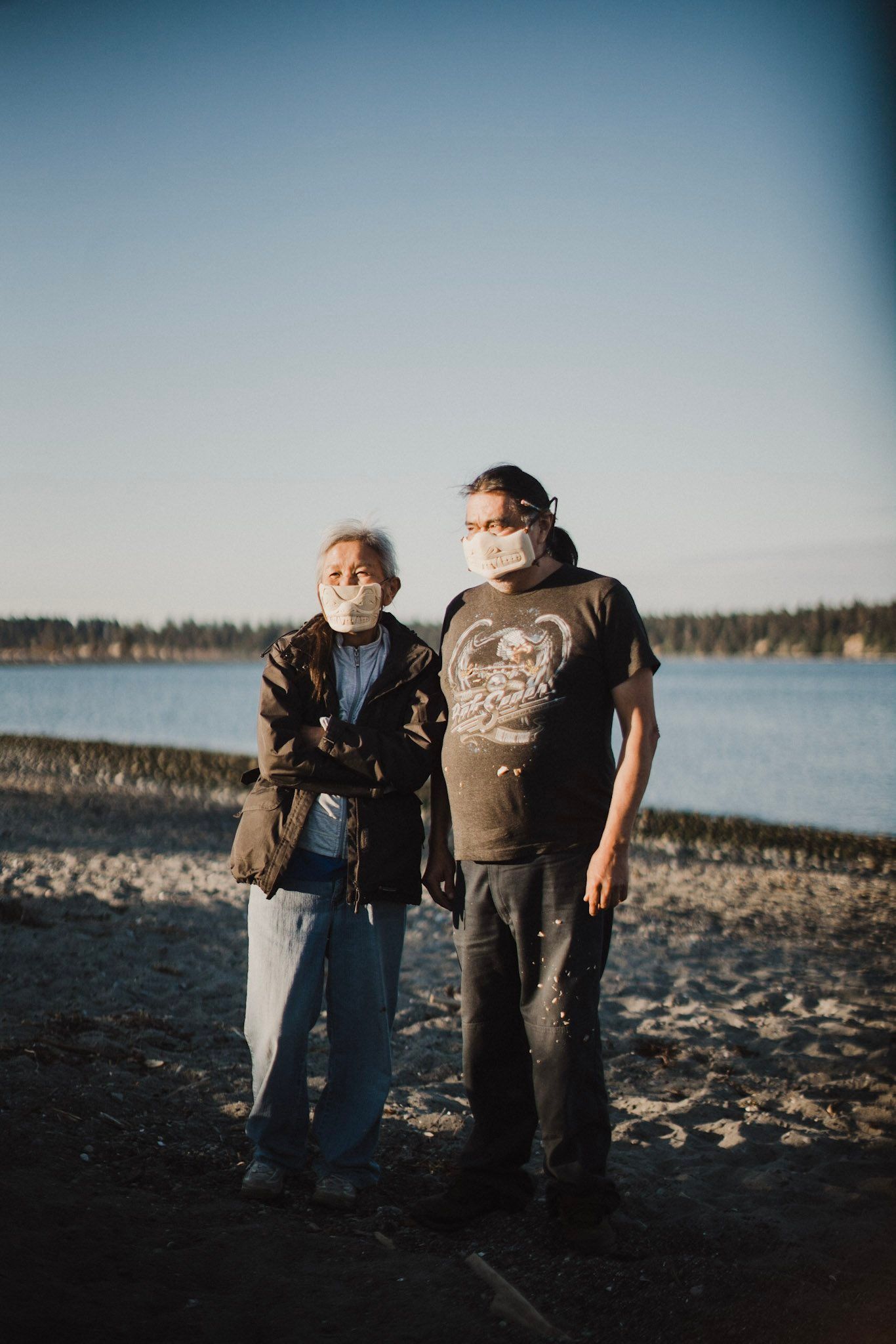 Two people stand on a rocky beach facing the camera, wearing masks, with forested shoreline and calm water behind them under clear sky