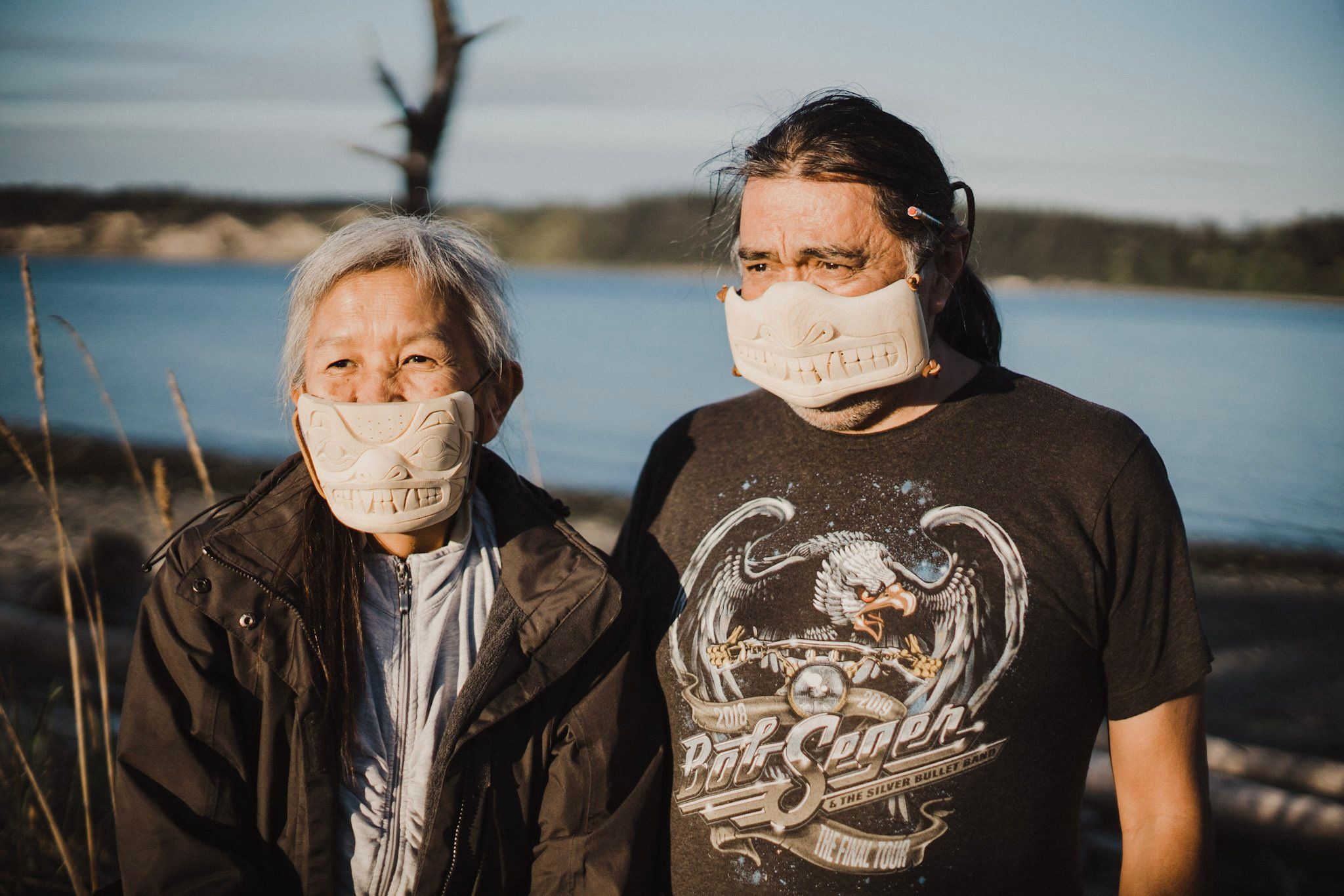 Two people wearing matching fabric masks stand by a calm blue lake under overcast sky, bare branches visible at left