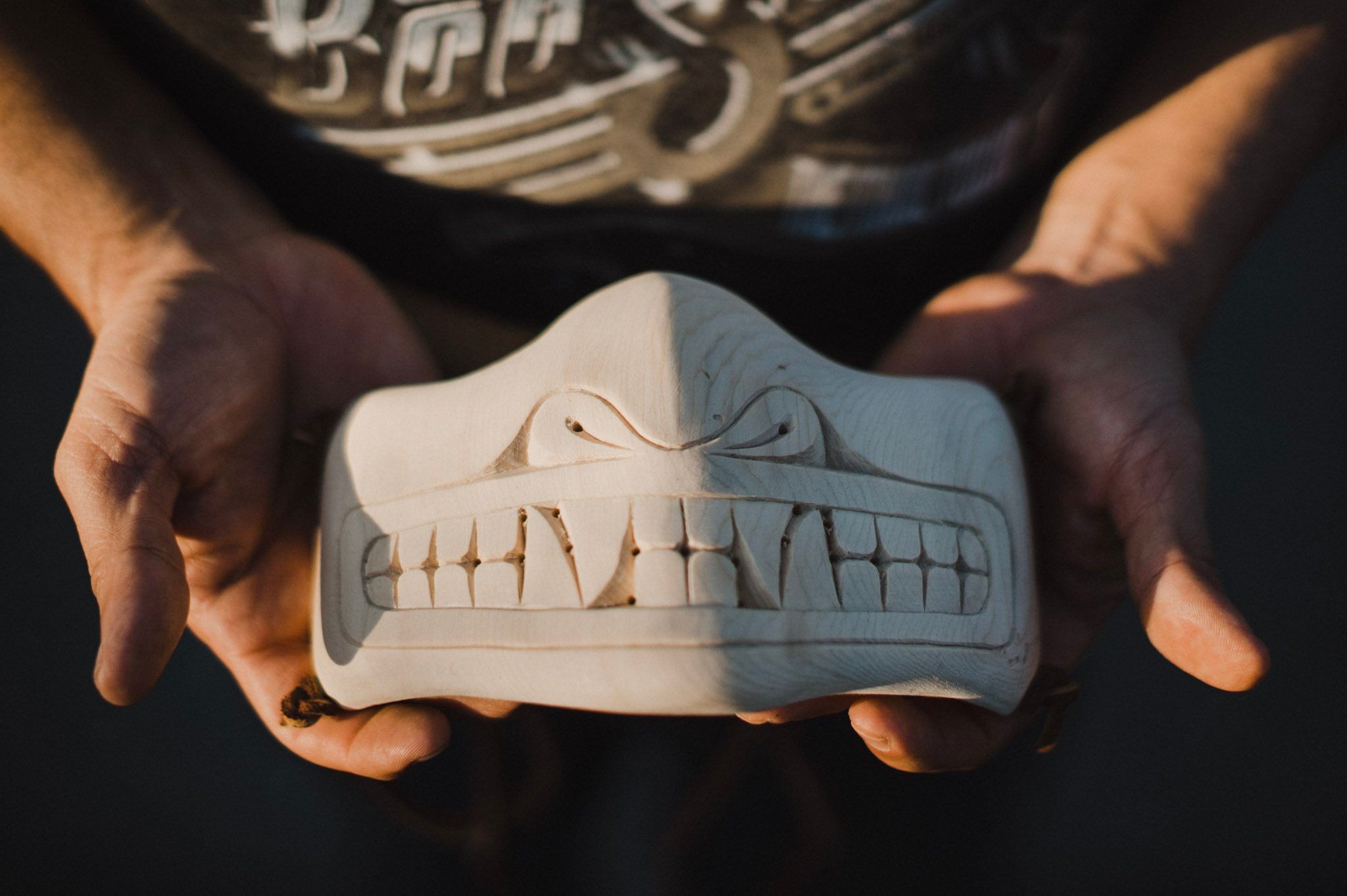 Hands hold a carved wooden mask showing two faces in profile against dark fabric, sunlight illuminating fine detail work