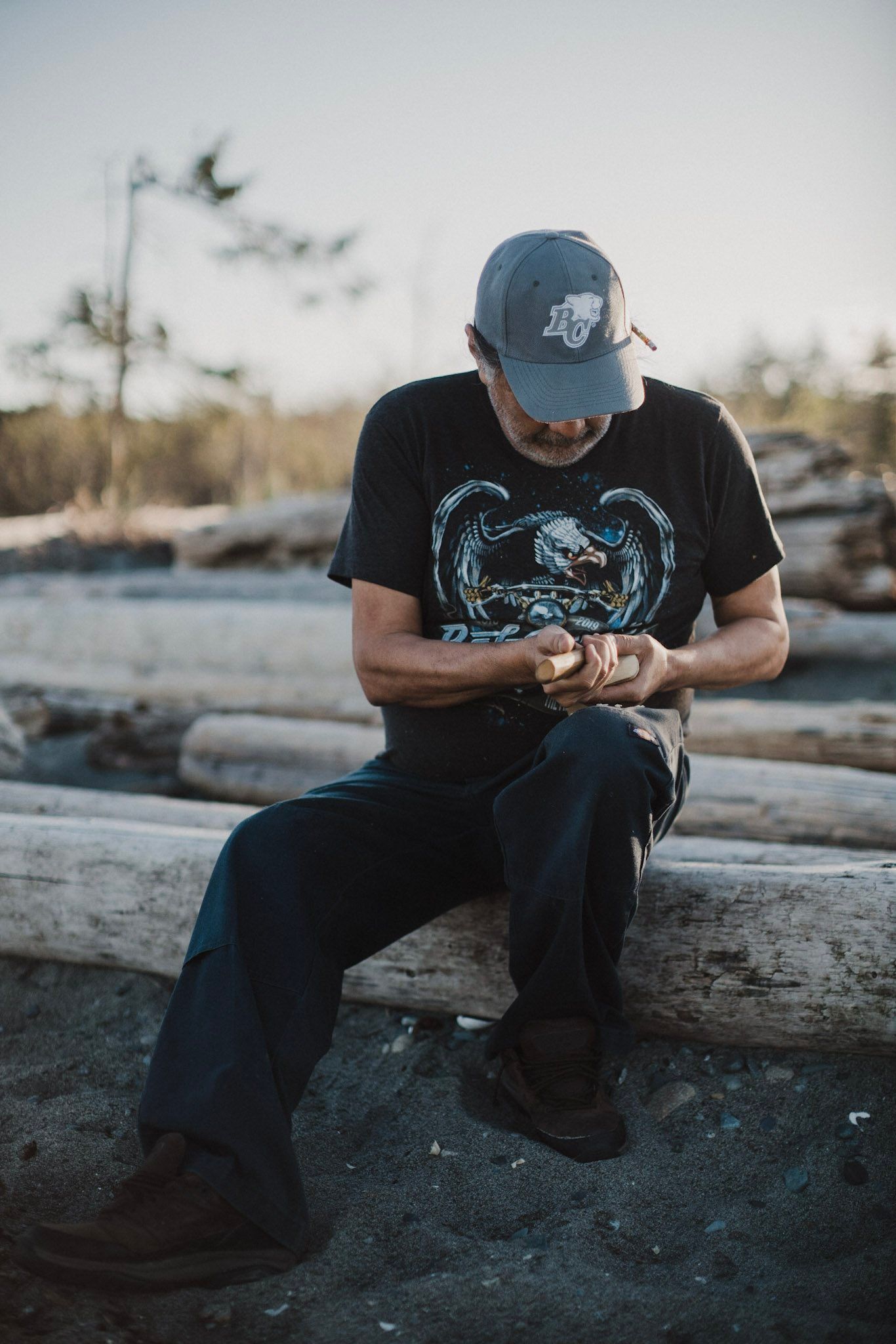 Man in cap and graphic tee sits on driftwood by water, studying his hands in golden late-day light