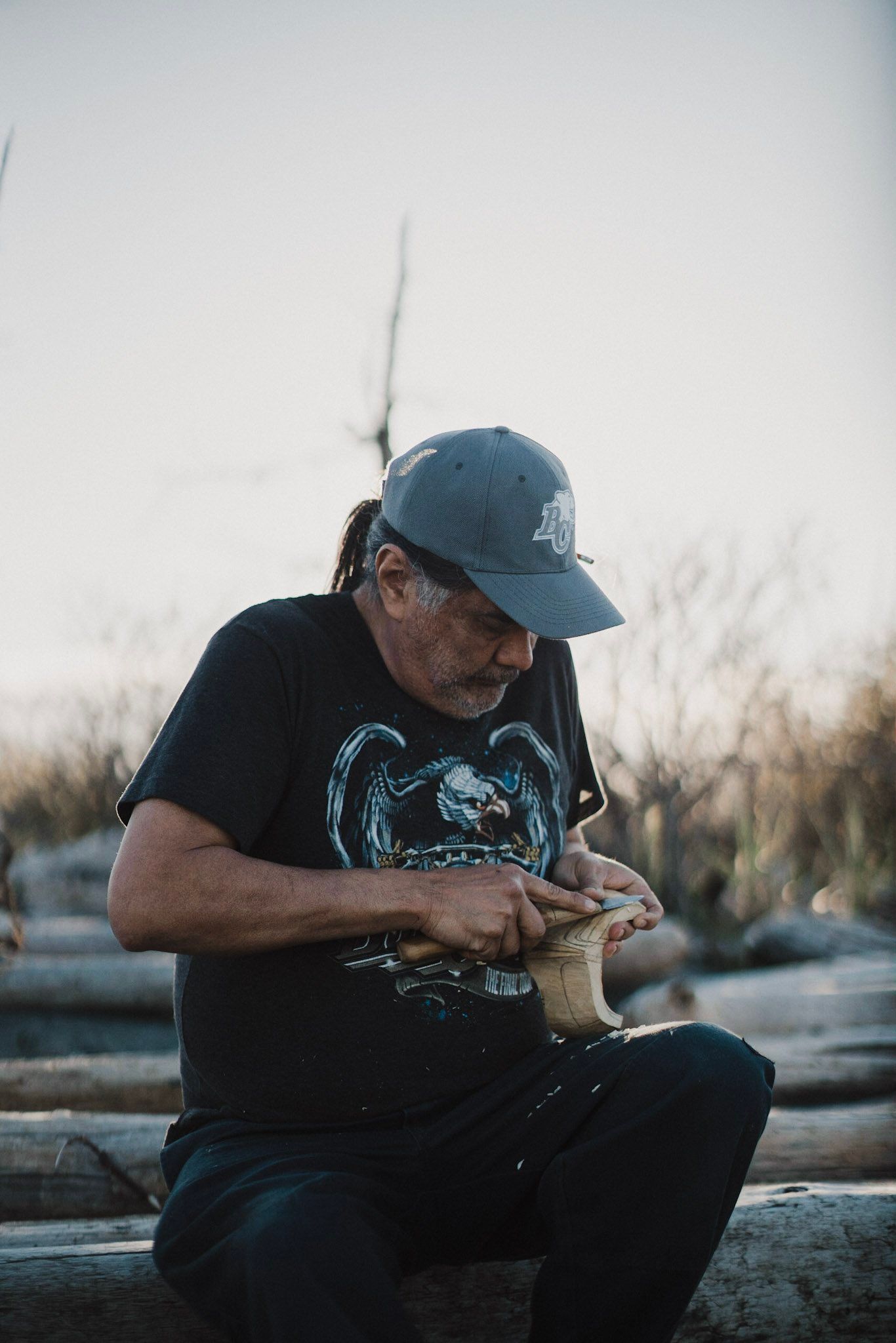 Man in blue cap examining carved wood detail while seated outdoors at dusk, misty forest backdrop