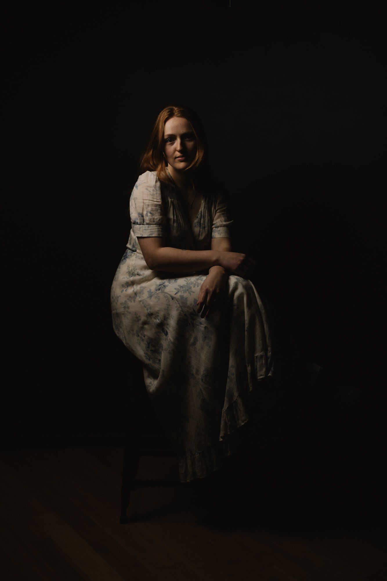 Woman in lace dress seated against dark background, warm spotlight illuminating her contemplative expression and delicate textile details