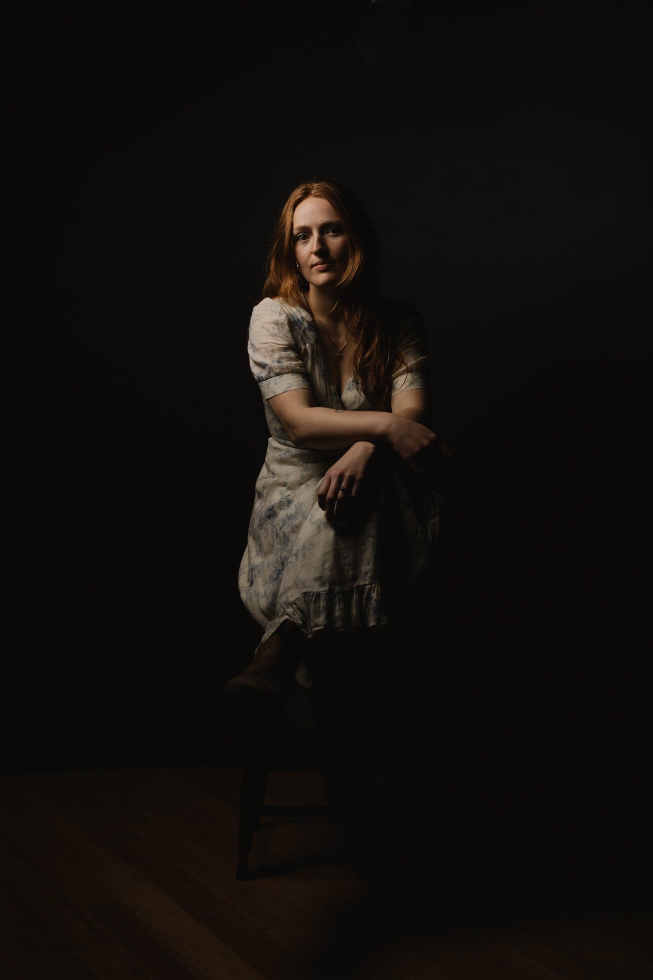 Woman in cream lace dress smiling against dark background, arms crossed, warm dramatic lighting on face and shoulders
