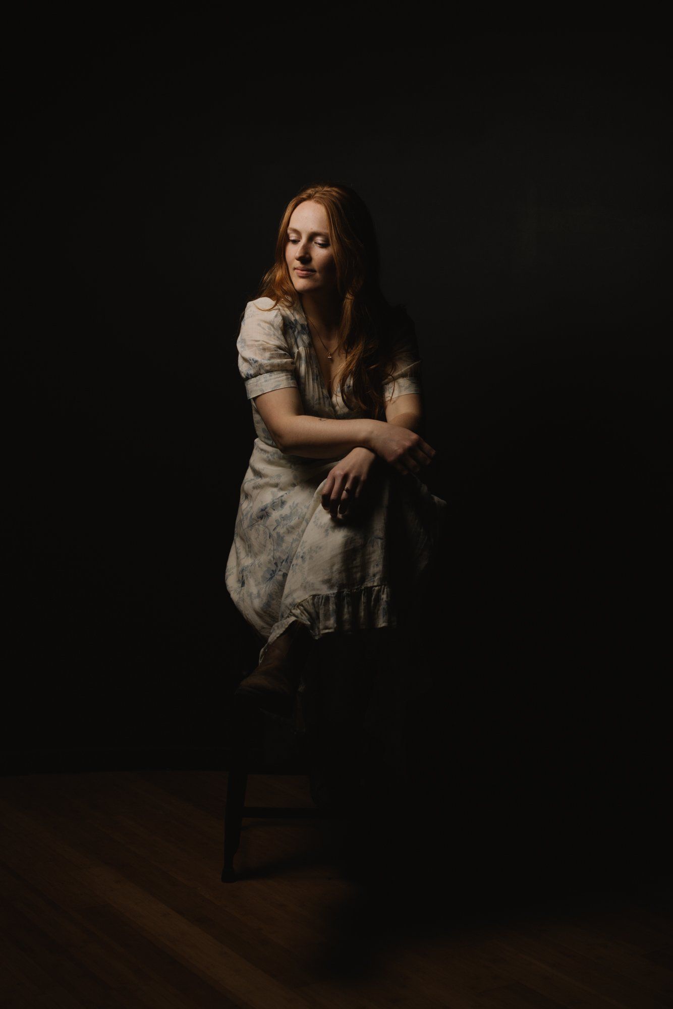 Woman in cream linen shirt seated against dark background, warm side lighting illuminating her contemplative gaze and gentle expression