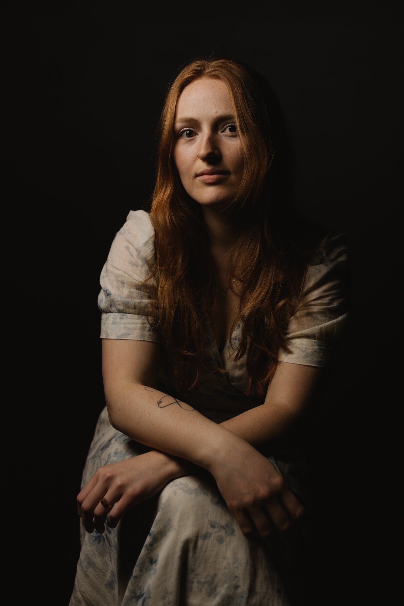 Woman with long reddish-brown hair in white linen shirt, arms crossed, gazing thoughtfully against dark background