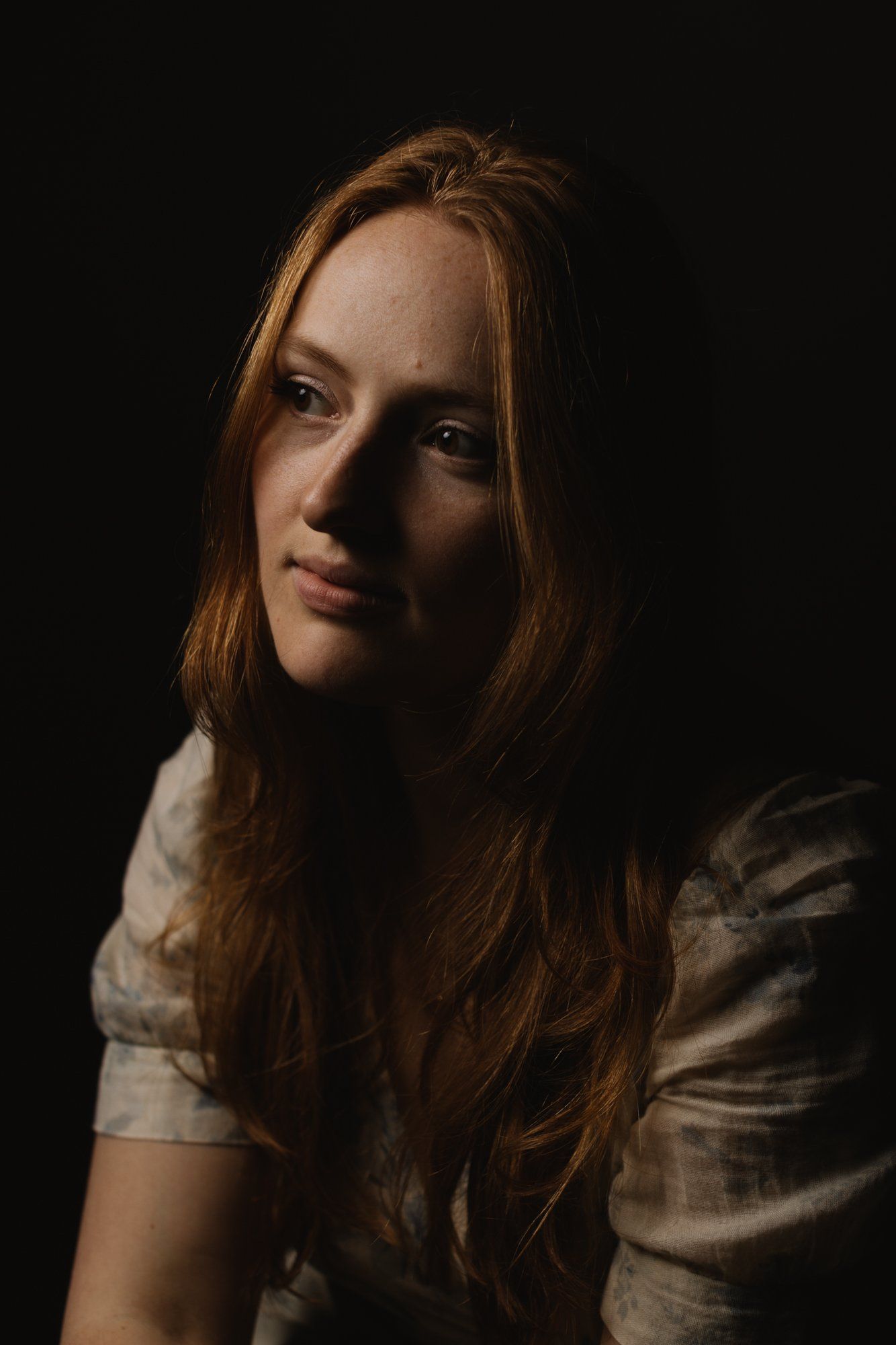 Young woman with long reddish-brown hair in soft white shirt, gazing toward camera against dark background with warm side lighting